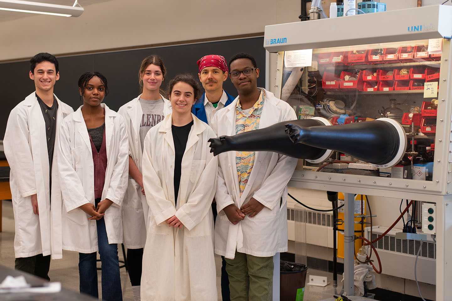A college professor in a chemistry lab with a group of students wearing white lab coats next to a glove box