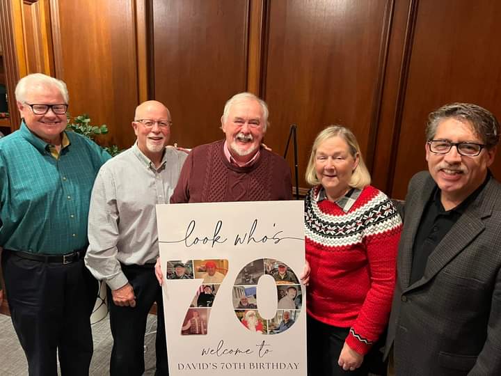 Five people pose together behind a sign that says "look who's 70"