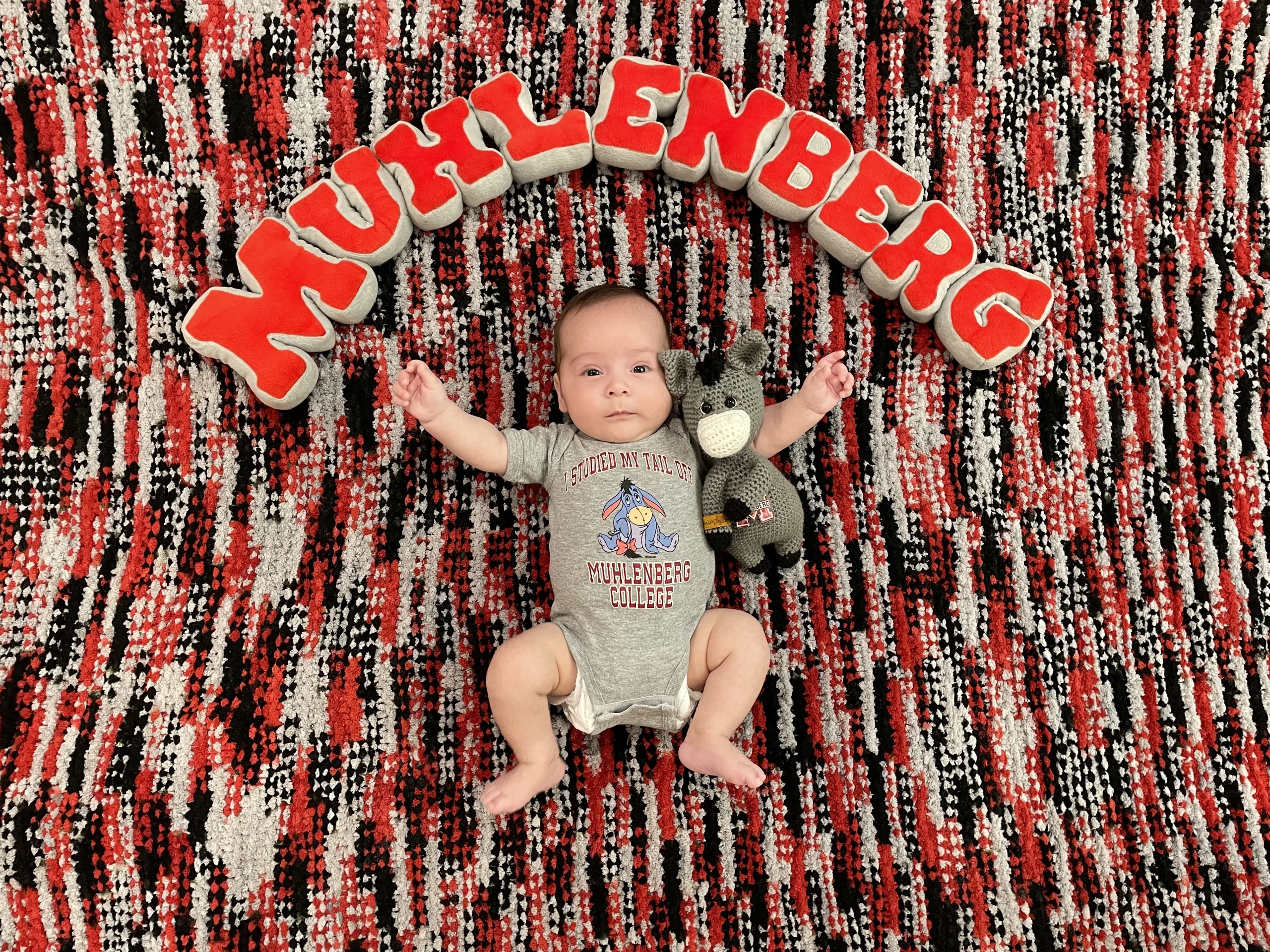 A baby in a Muhlenberg onesie lays on a red, black, and gray blanket under stuffed letters that read MUHLENBERG