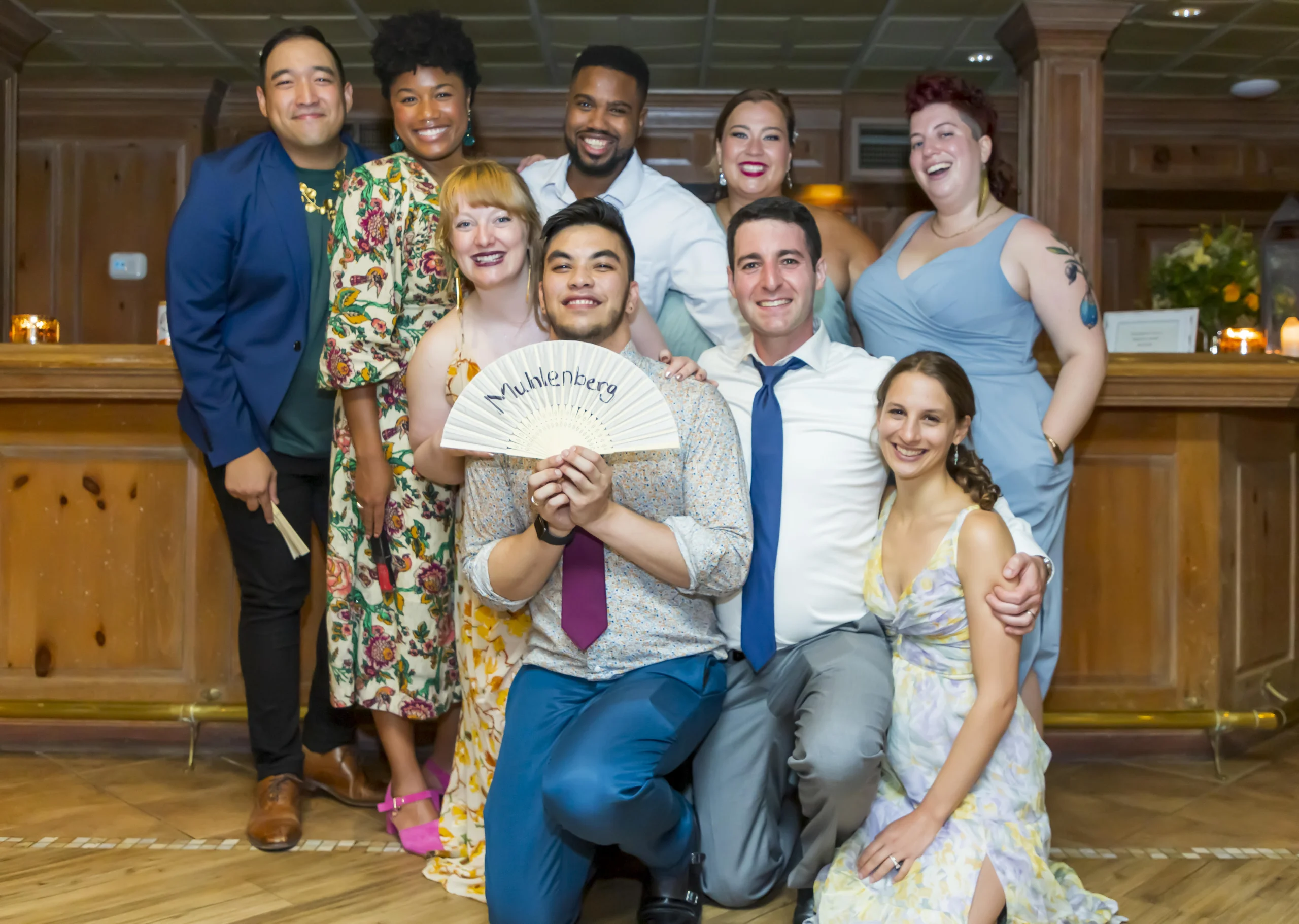 A group of friends at a wedding, including the two grooms, pose with a fan that reads "Muhlenberg"