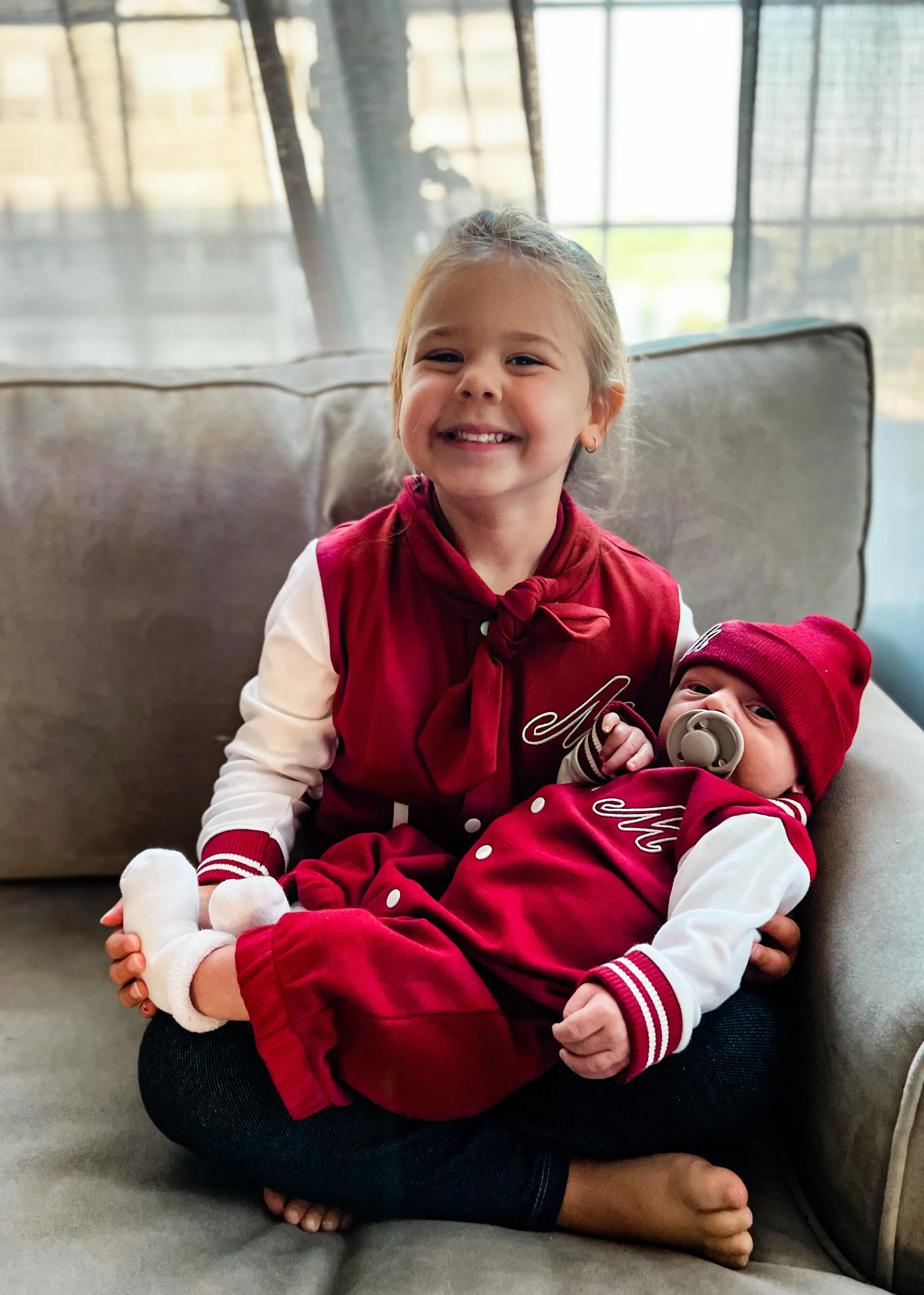 A little girl holds a baby; both are wearing red jackets with a script M on them