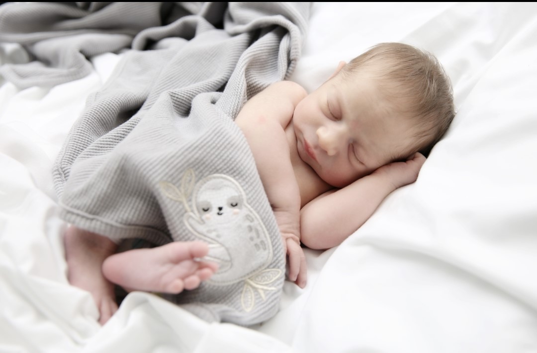 A baby sleeps on a white sheet beneath a gray blanket