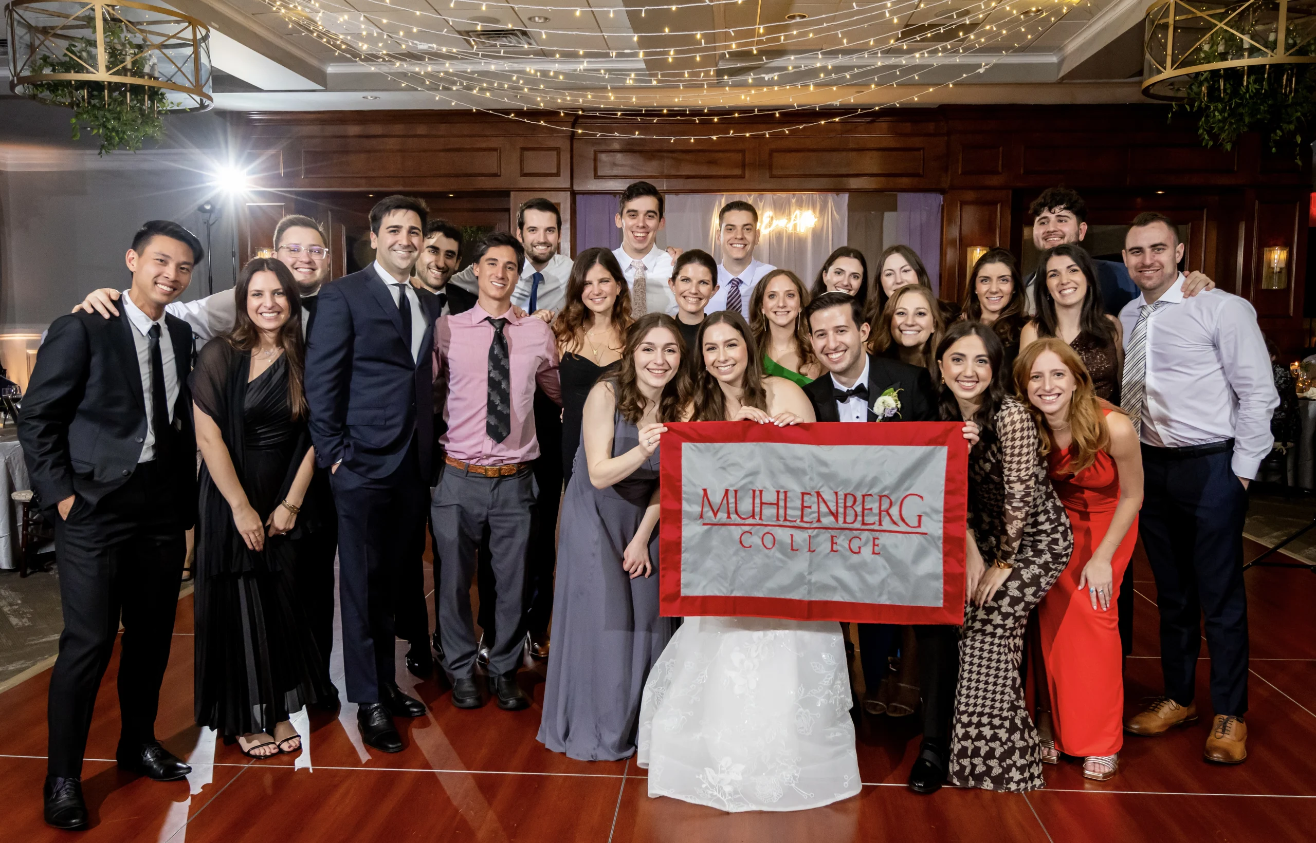 At a wedding reception, the bride and groom and friends hold a flag that says Muhlenberg College