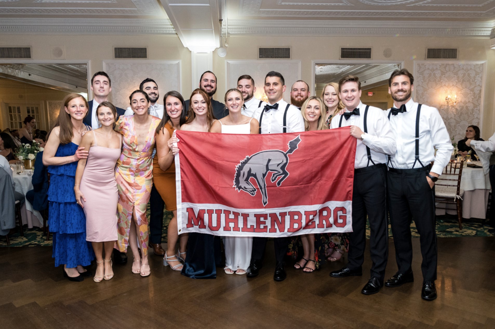 At a wedding reception, the bride and groom and friends hold a flag that says Muhlenberg