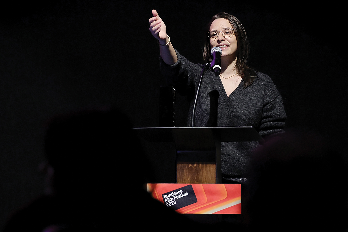A woman wearing glasses speaks at a podium and raises her hand toward the audience, a placard on the podium reads Sundance Film Festival 2023