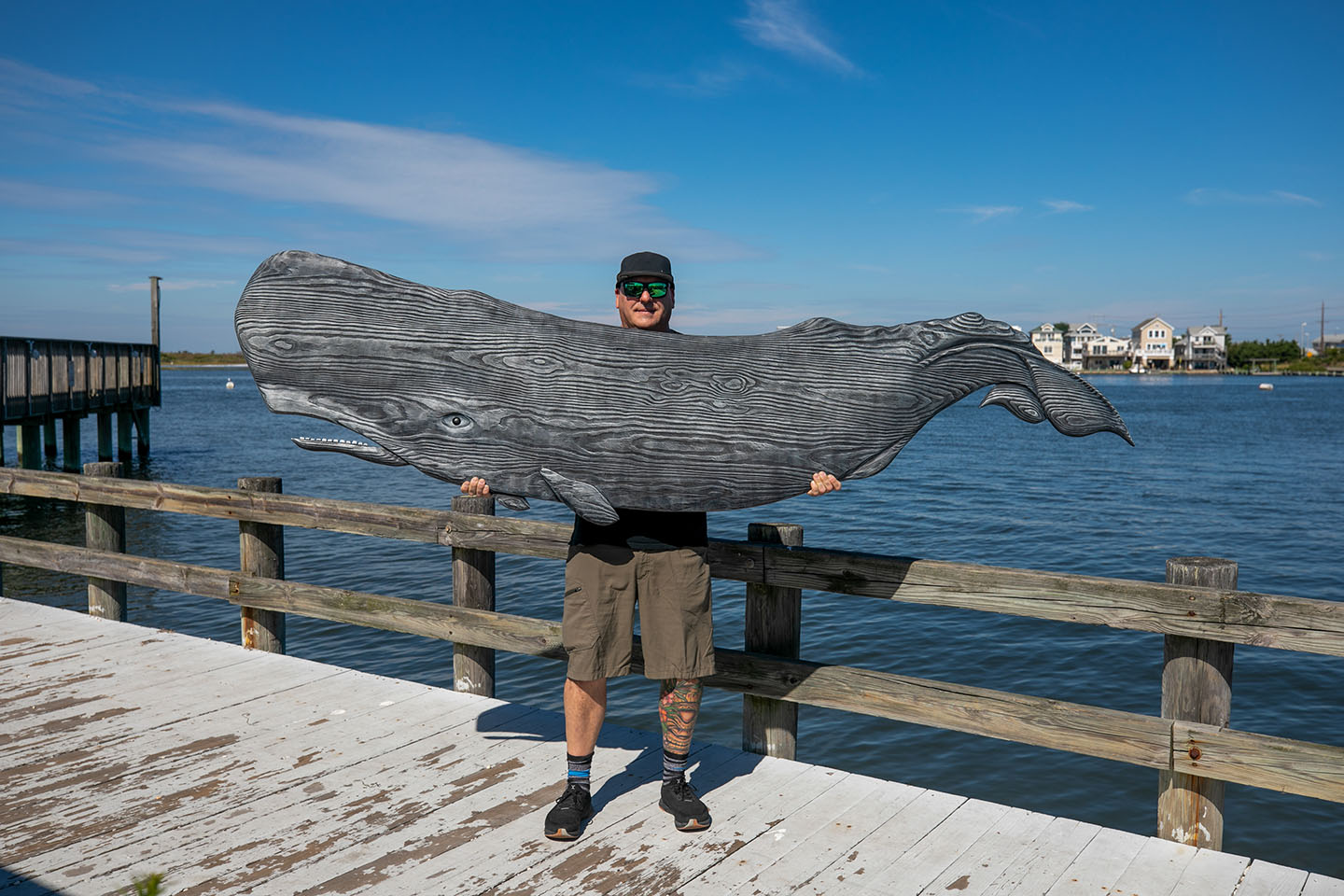 A man in a baseball hat holds a massive carved wooden whale while standing on a pier