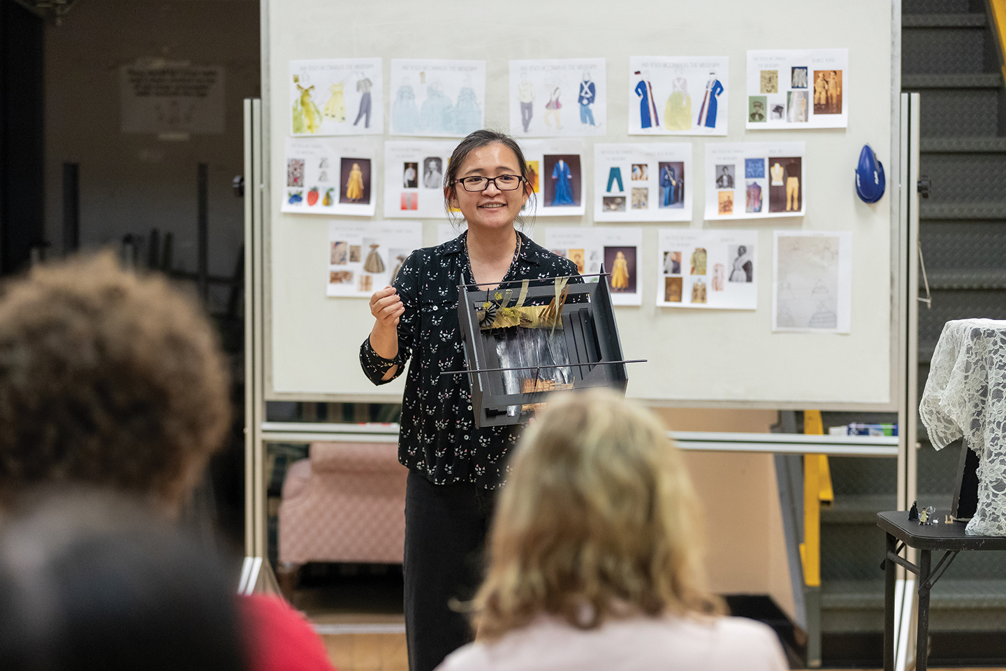 A college professor stands in front of a room of students and shows them a set model for a play