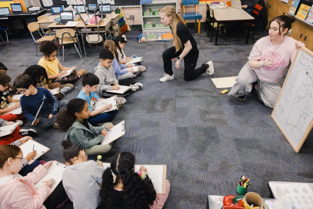 Second graders sit on the floor and use clipboards to draw