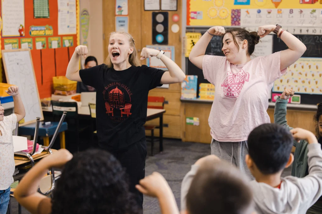 Two college students flex their arms and speak to a group of second graders who are doing the same