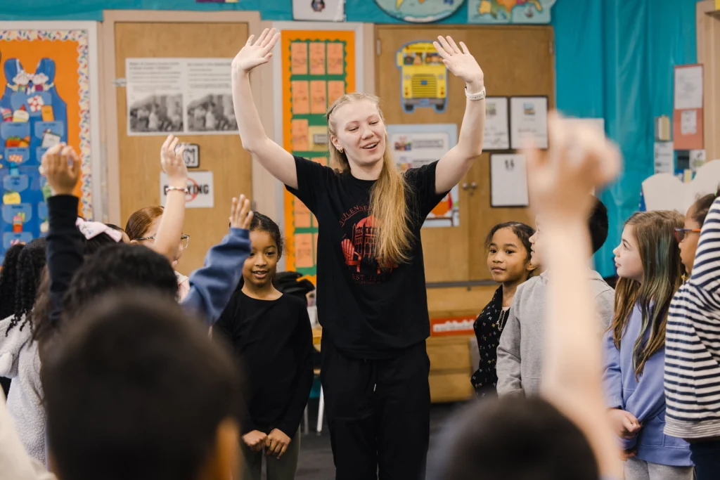 A college student stands among second graders with her arms up in the air and while smiling