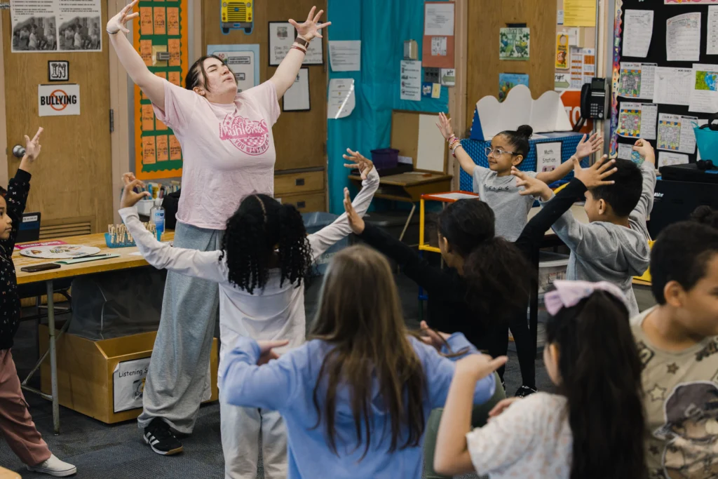 A college student makes her arms really big as she leads second graders in a dance class
