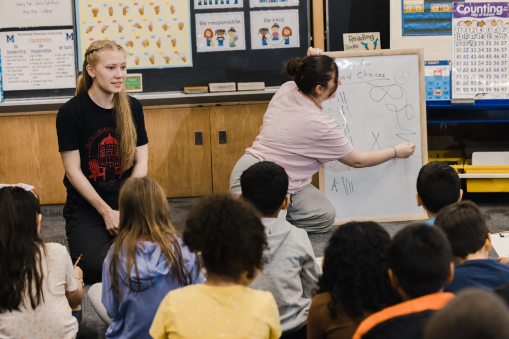 College students use a whiteboard to show second graders how to draw a treasure map