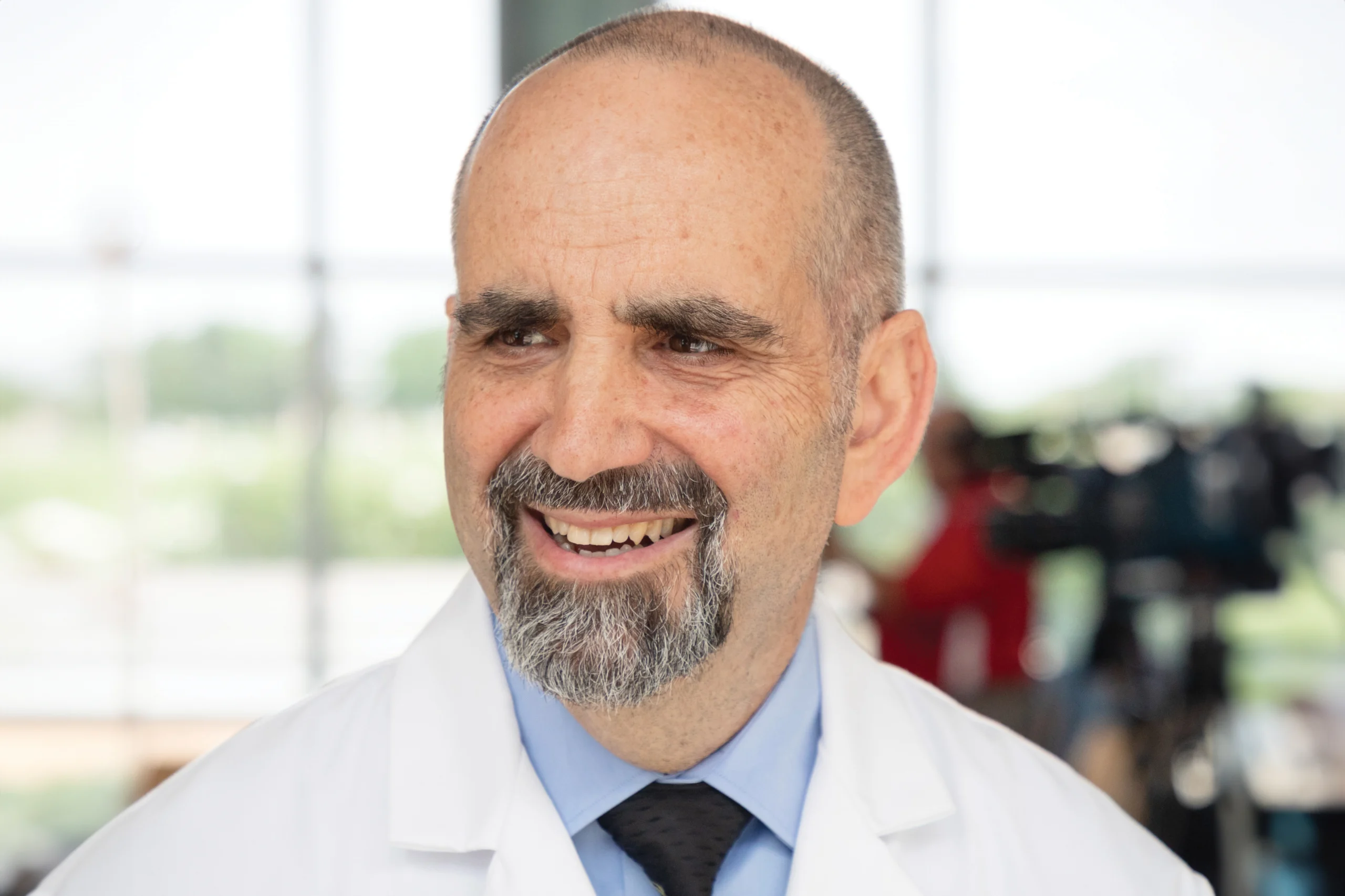 Portrait photo of an older man in a lab coat and blue collared shirt with black tie smiling for the camera while looking off to the left