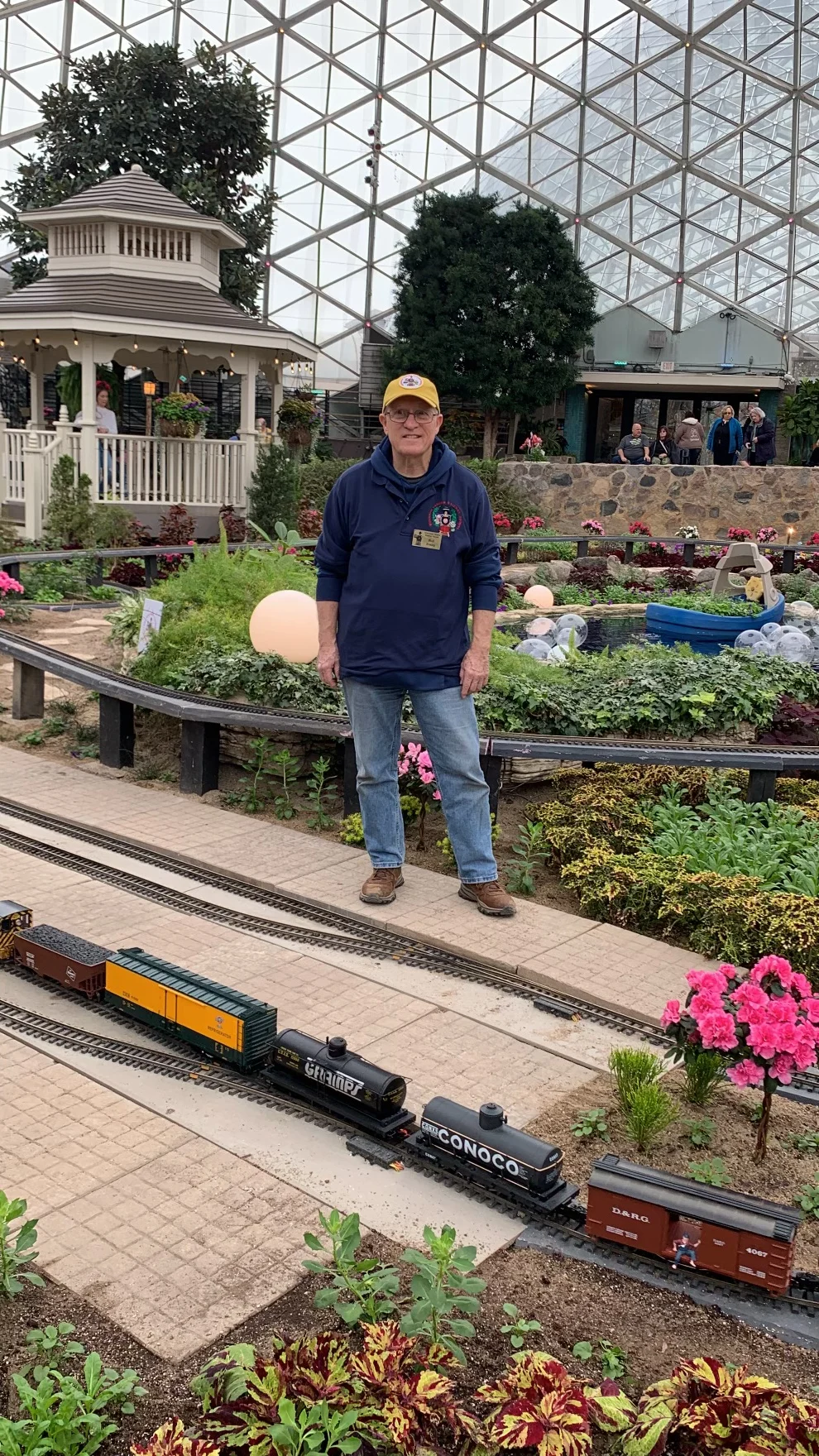 An older man in a blue sweatshirt and yellow hat standing in the middle of a greenhouse harden with model train running.