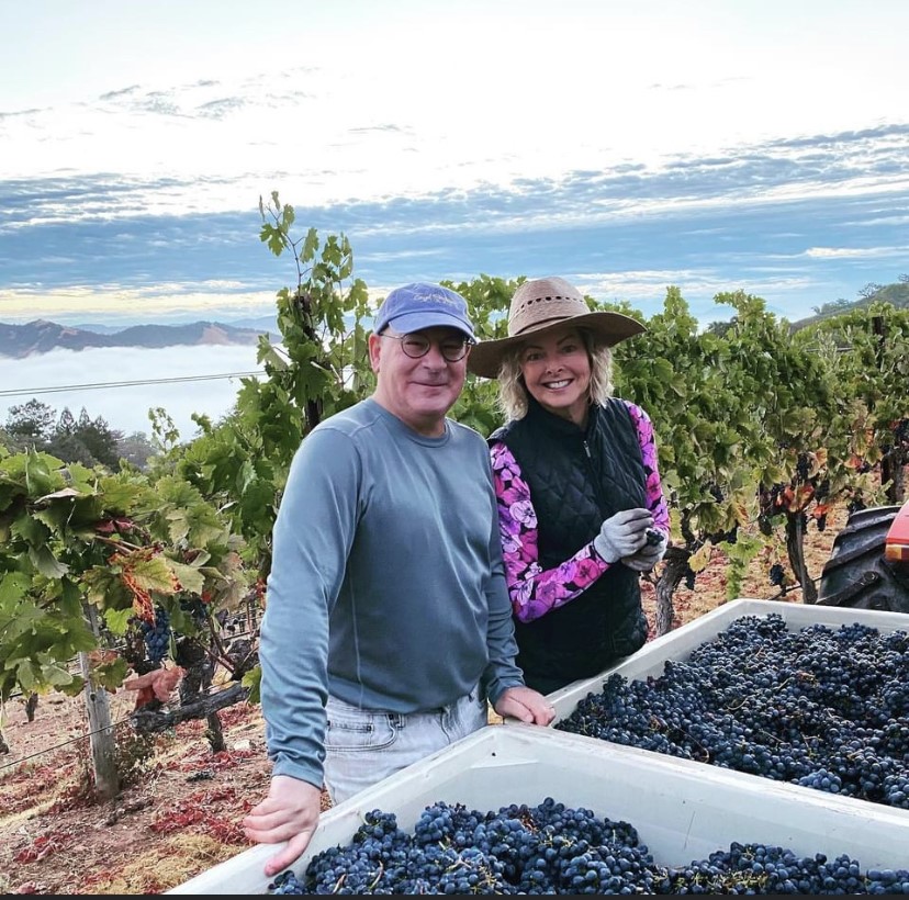 Two people in hats smile for a picture in a vineyard with bins of grapes in front of them