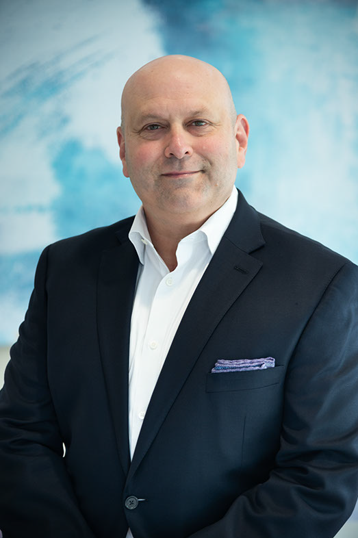 A headshot of a man in a dark blue suit with a light blue background