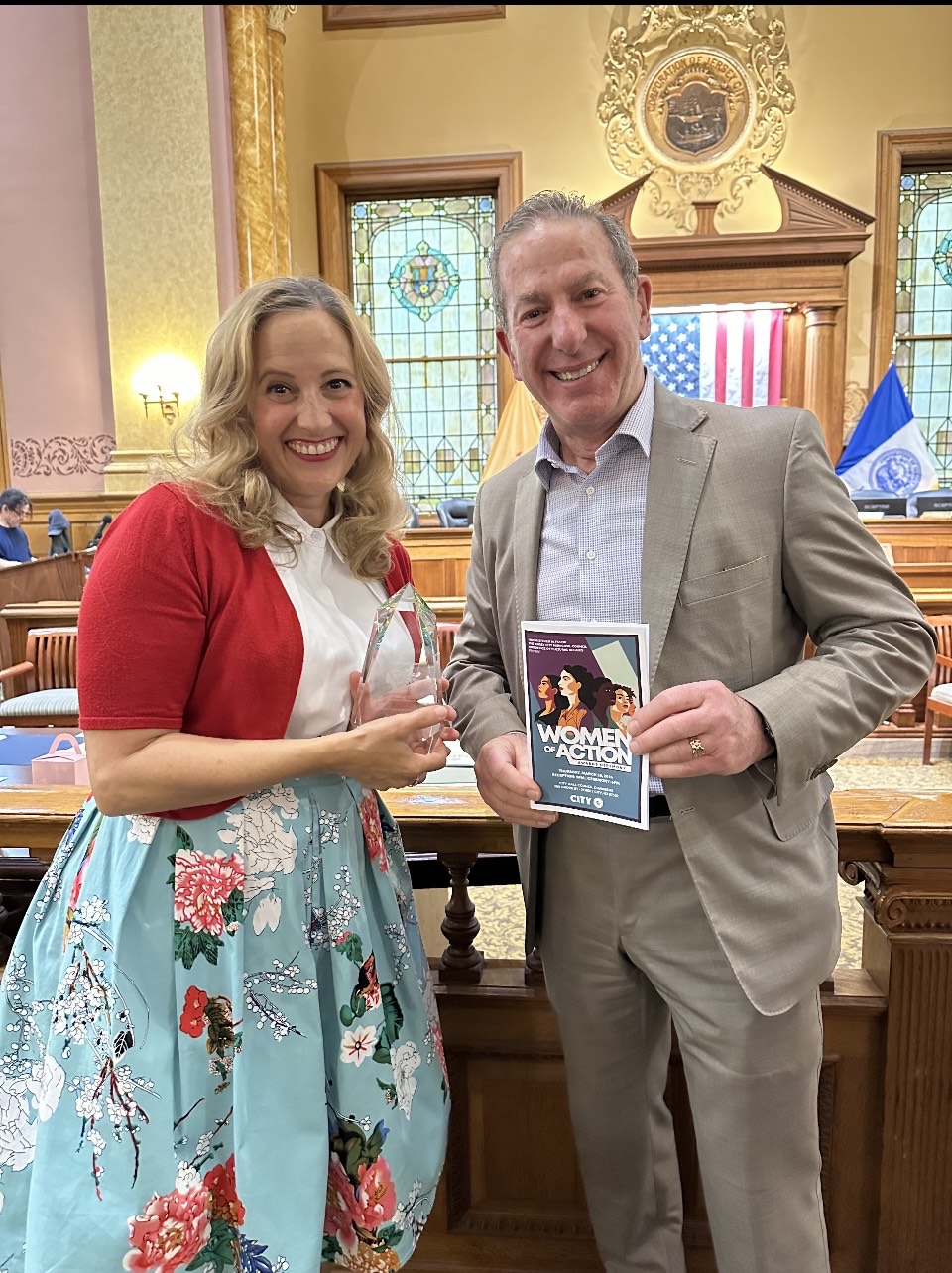 Two people smile for a picture in a government building, the one on the left holds an award