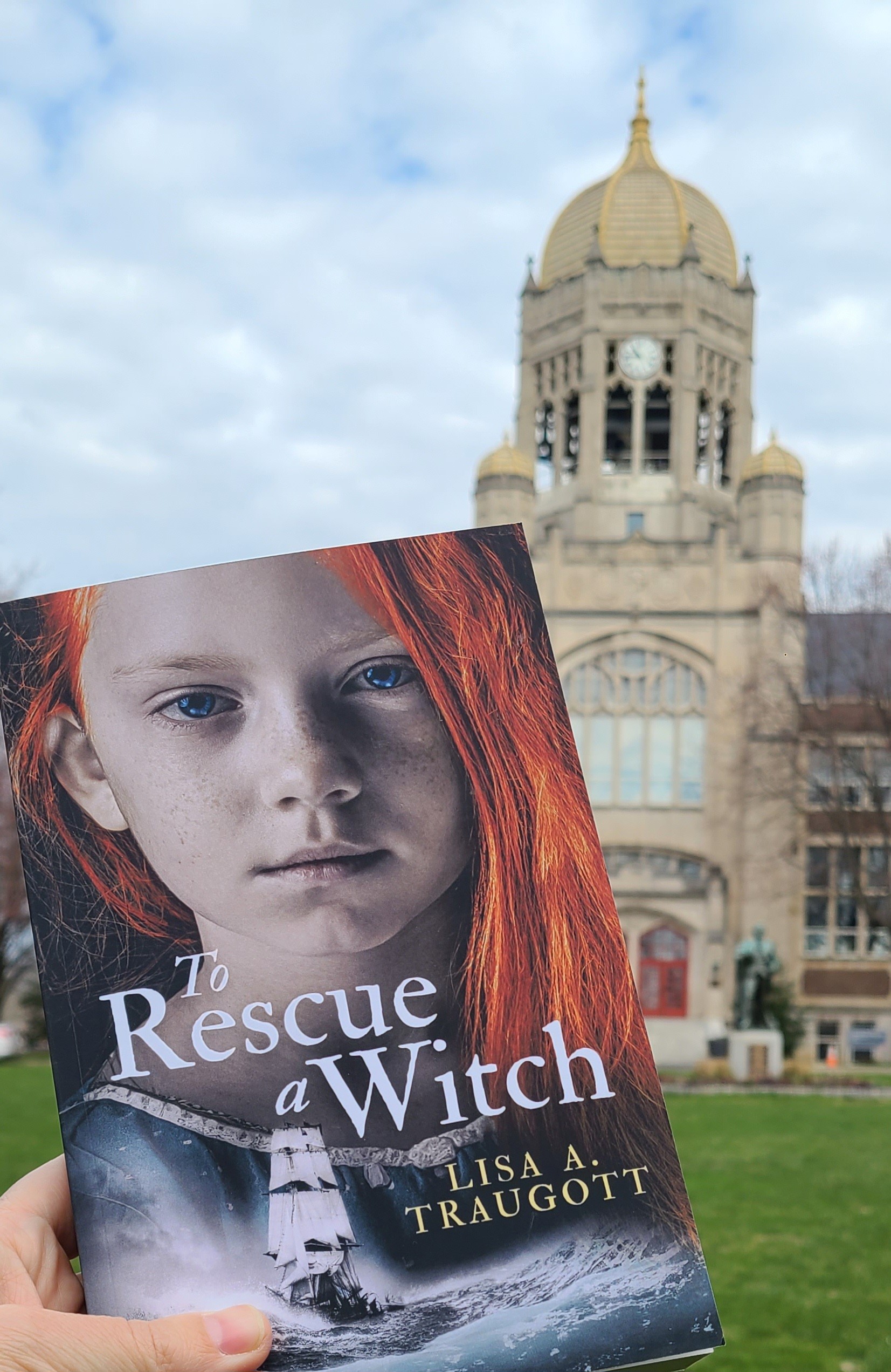 A hand holds up a book with a cover that says To Rescue a Witch in front of the Haas Bell Tower on Muhlenberg's campus