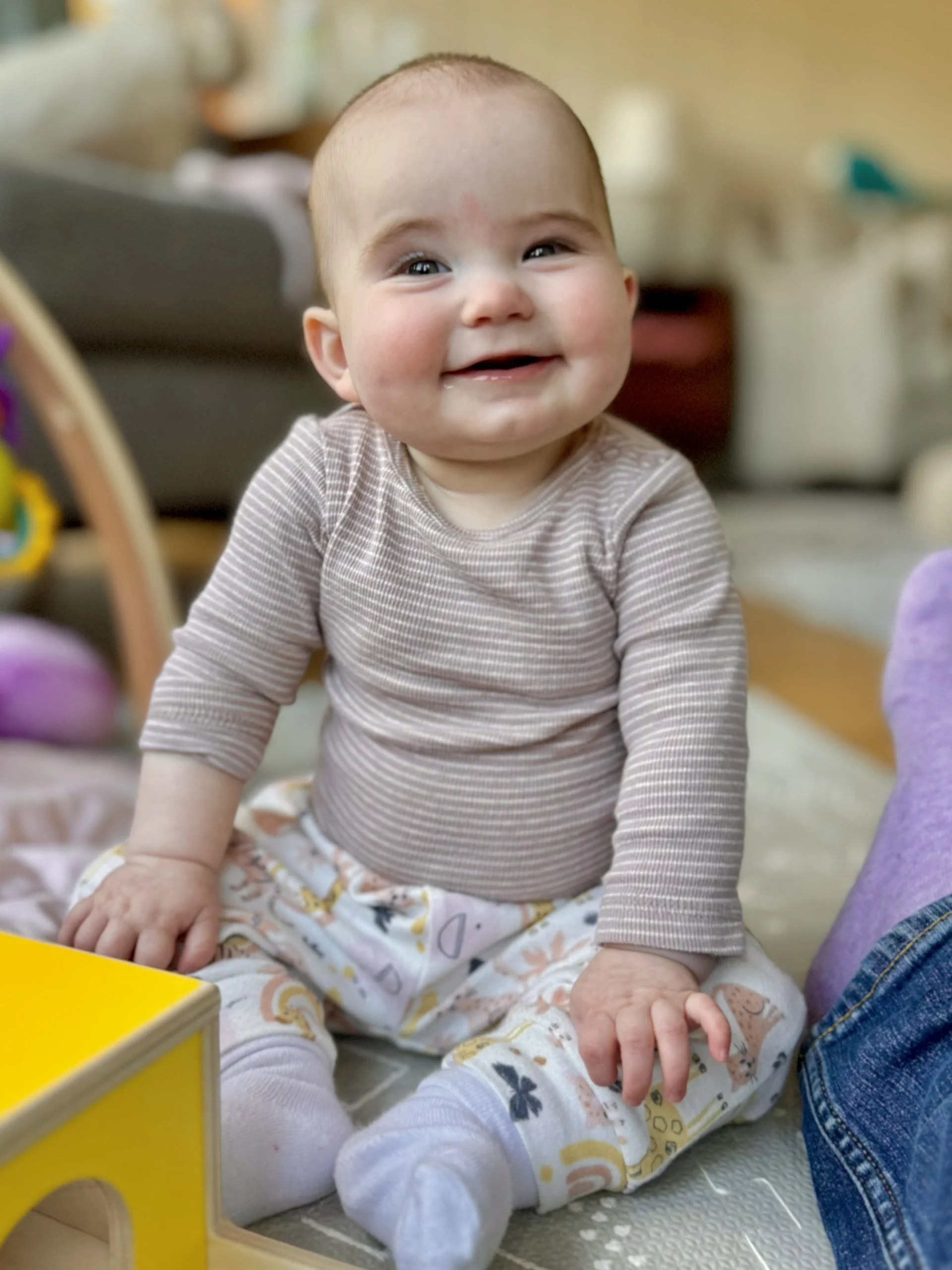 A smiling baby sitting on the floor in a brown striped shirt and white pants with animals on them