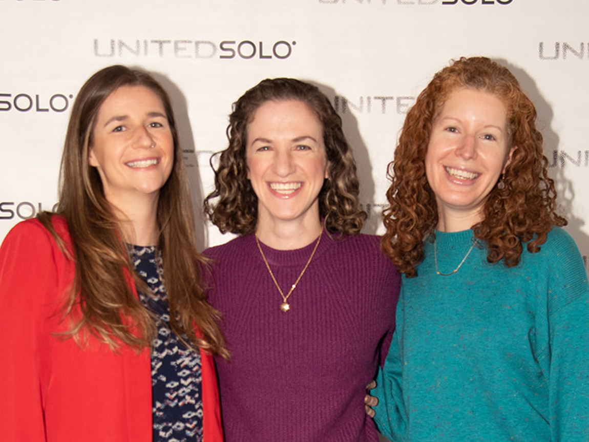 Three people with long hair smile for a photo in front of a backdrop that says UNITEDSOLO