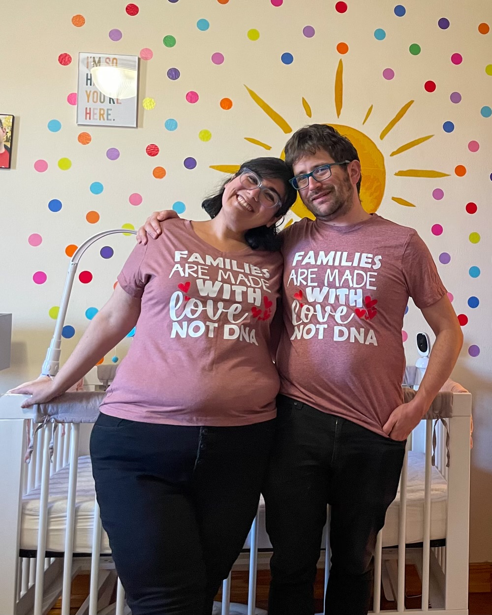 A couple wearing matching shirts that say "FAMILIES ARE MADE WITH LOVE NOT DNA" stand in front of a crib and a colorful wall mural