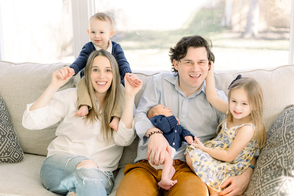 A family with three young children, including an infant, smiles for a photo on a beige couch