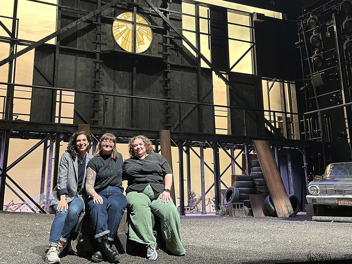 Three people smile for a photo on the set of a Broadway musical