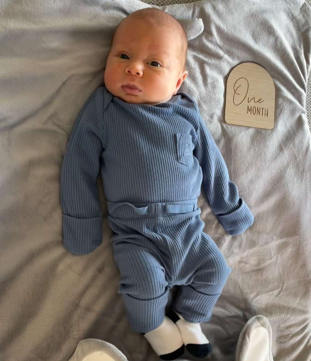 A baby in a blue outfit reclines next to a placard that says One Month