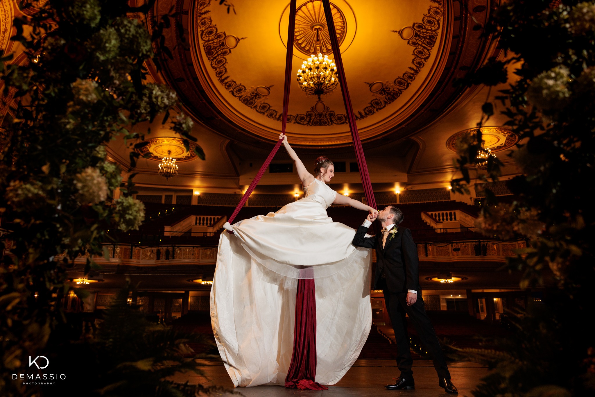 A woman in a wedding dress does a split while hanging from aerial silks in a theatre while the groom kisses her hand