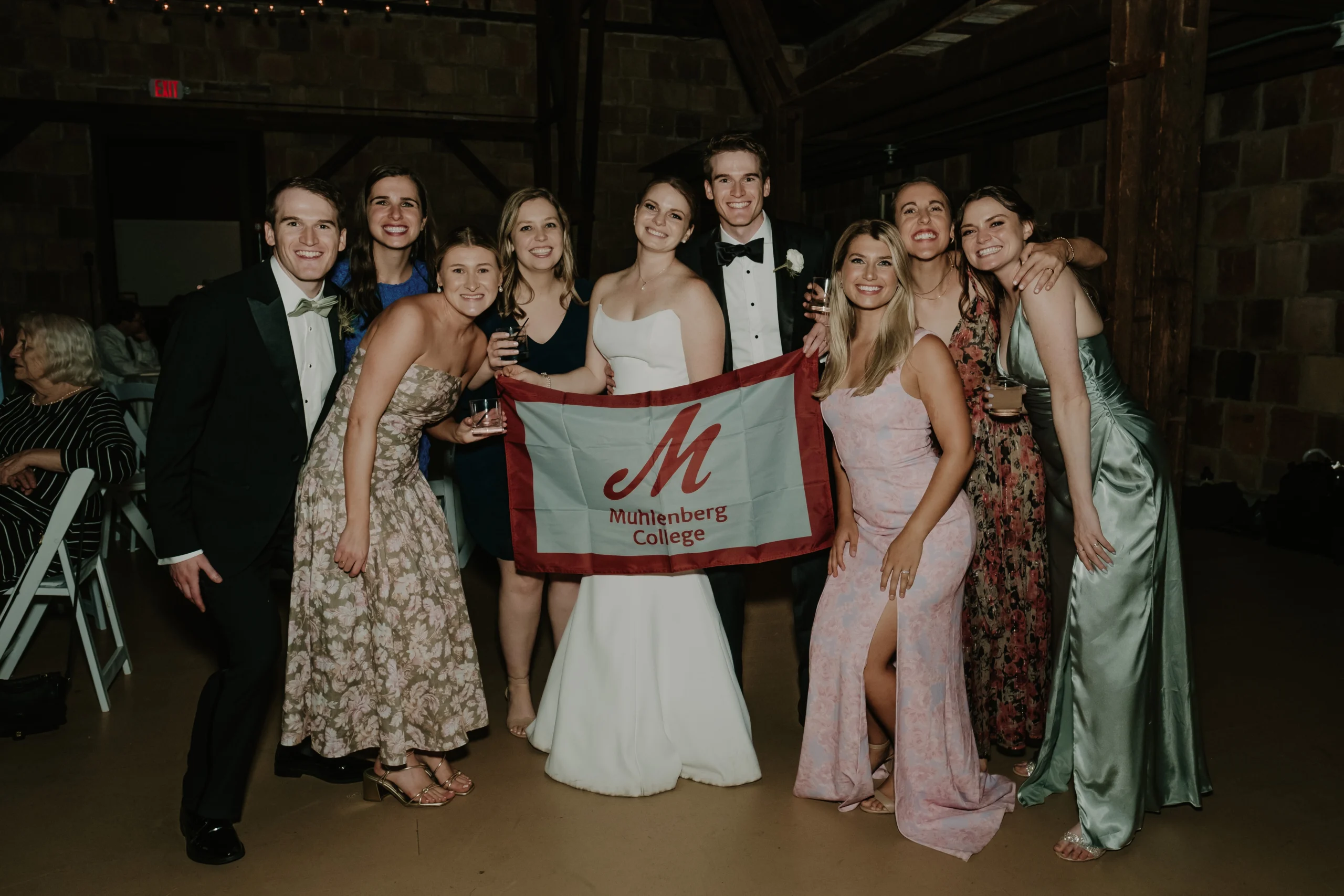 A group of young people gathered together to smile for a photo while holding a red trimmed gray flag with a red M and Muhlenberg College written on it