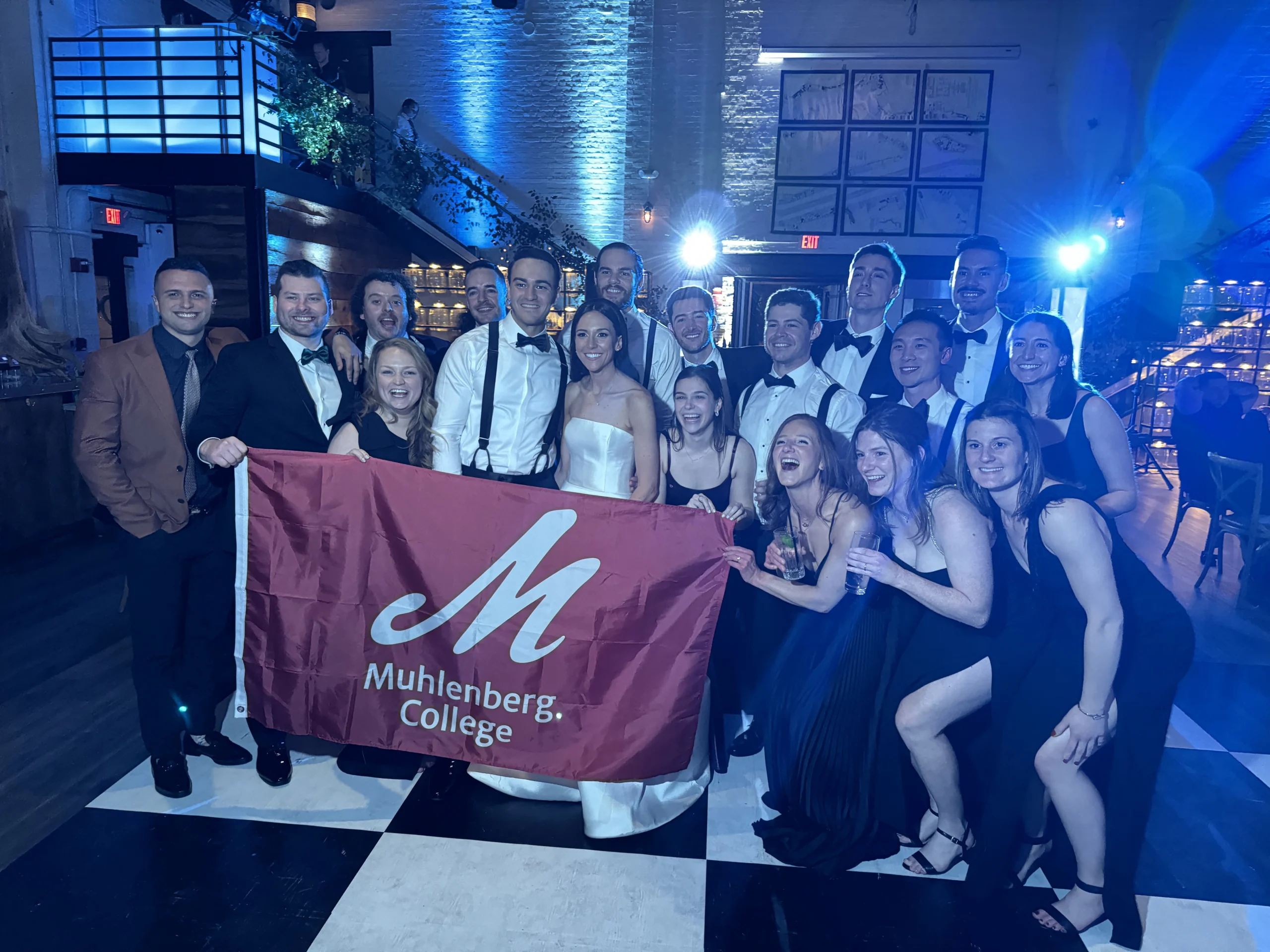 A group of young people gathered together to smile for a photo while holding a red flag with a white M and Muhlenberg College written on it