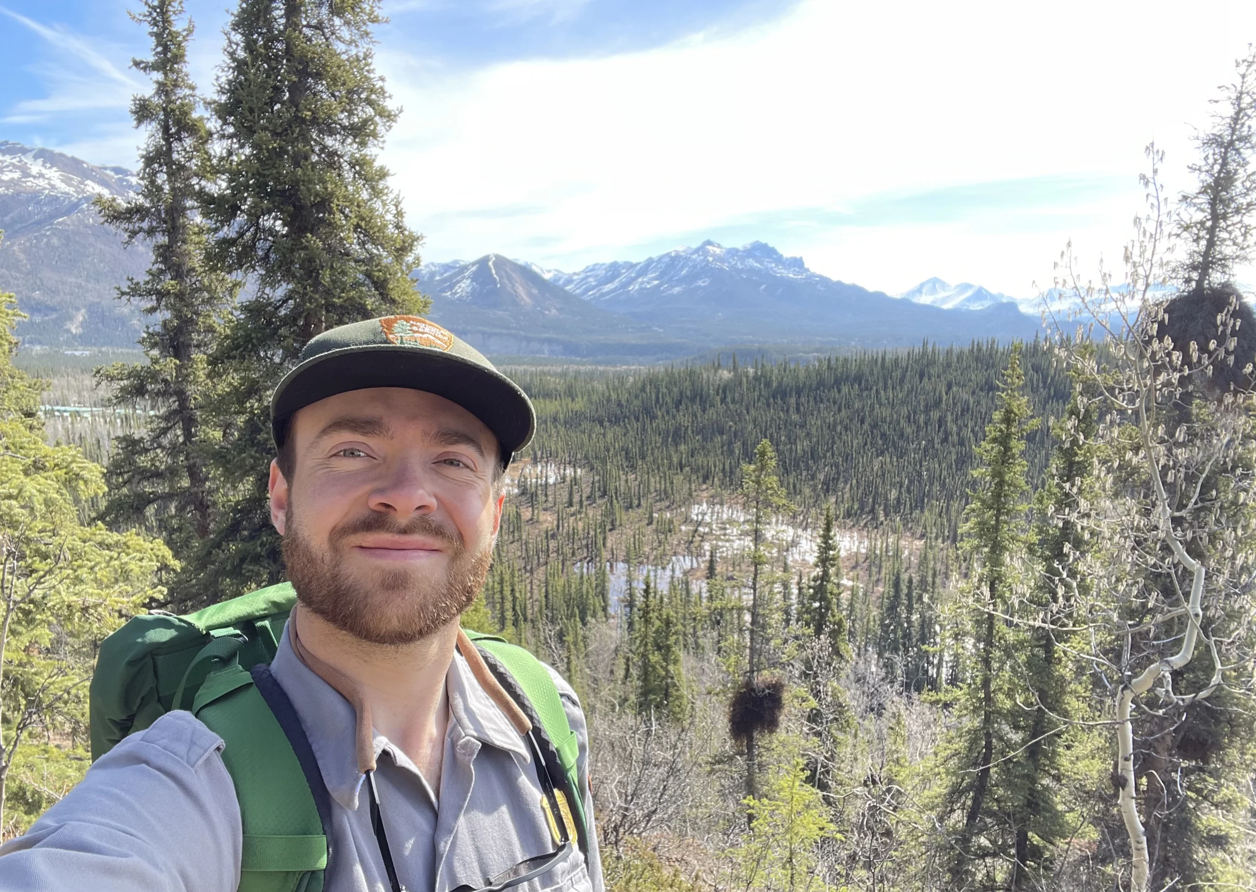 A young man takes a selfie in the woods with mountains in the background while wearing a green backpack, green hat, and light gray shirt.