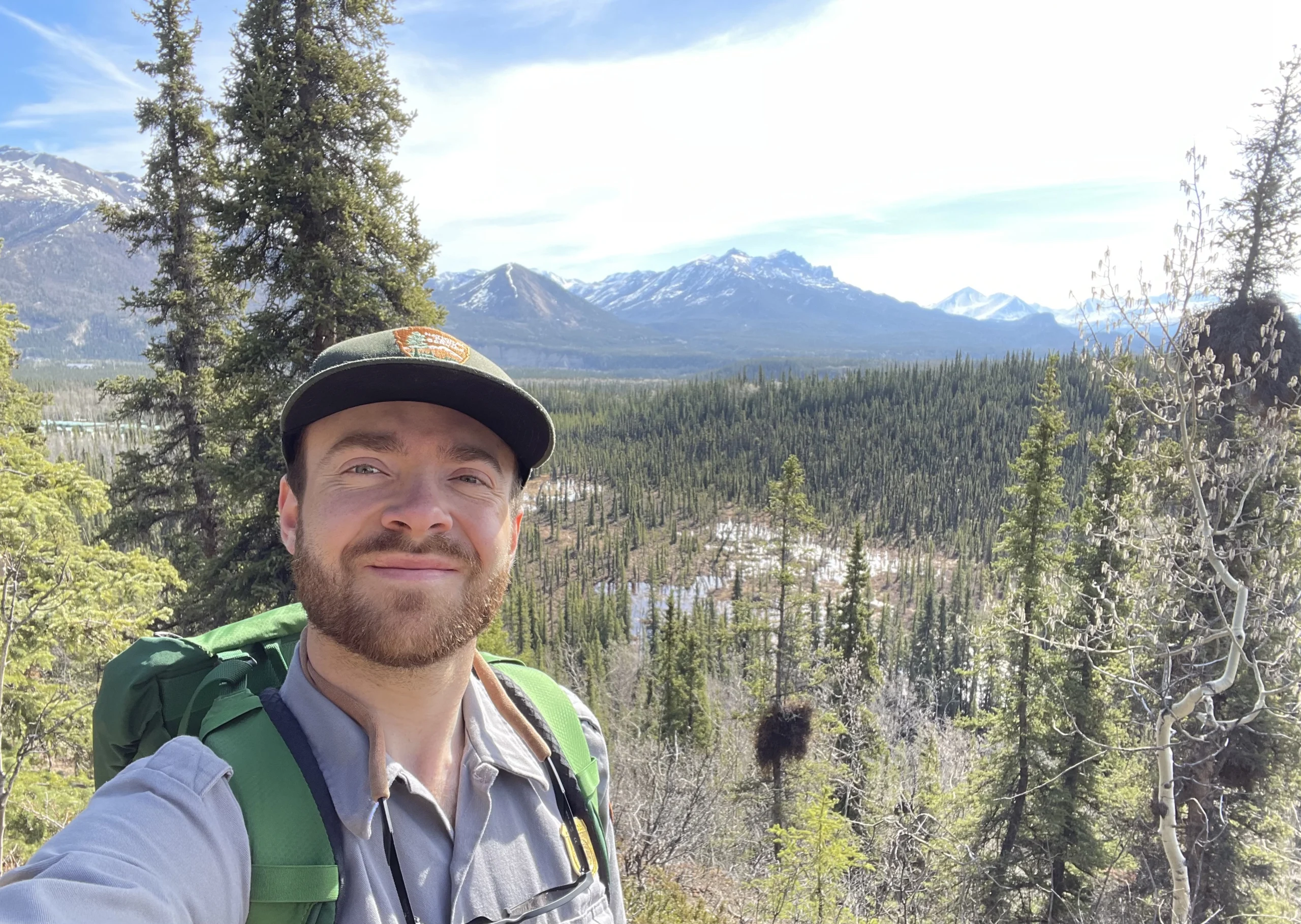 A young adult in a hat with a beard takes a selfie in the forest with snow-capped mountains in the background