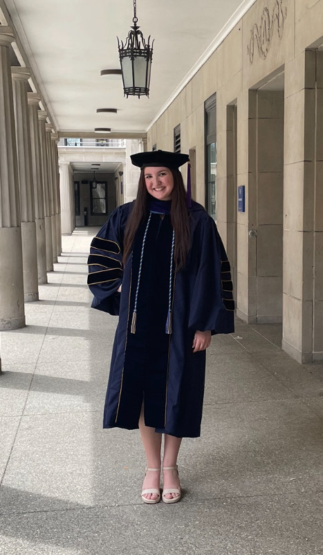 A young adult with long dark hair poses for a photo in graduation regalia for law school