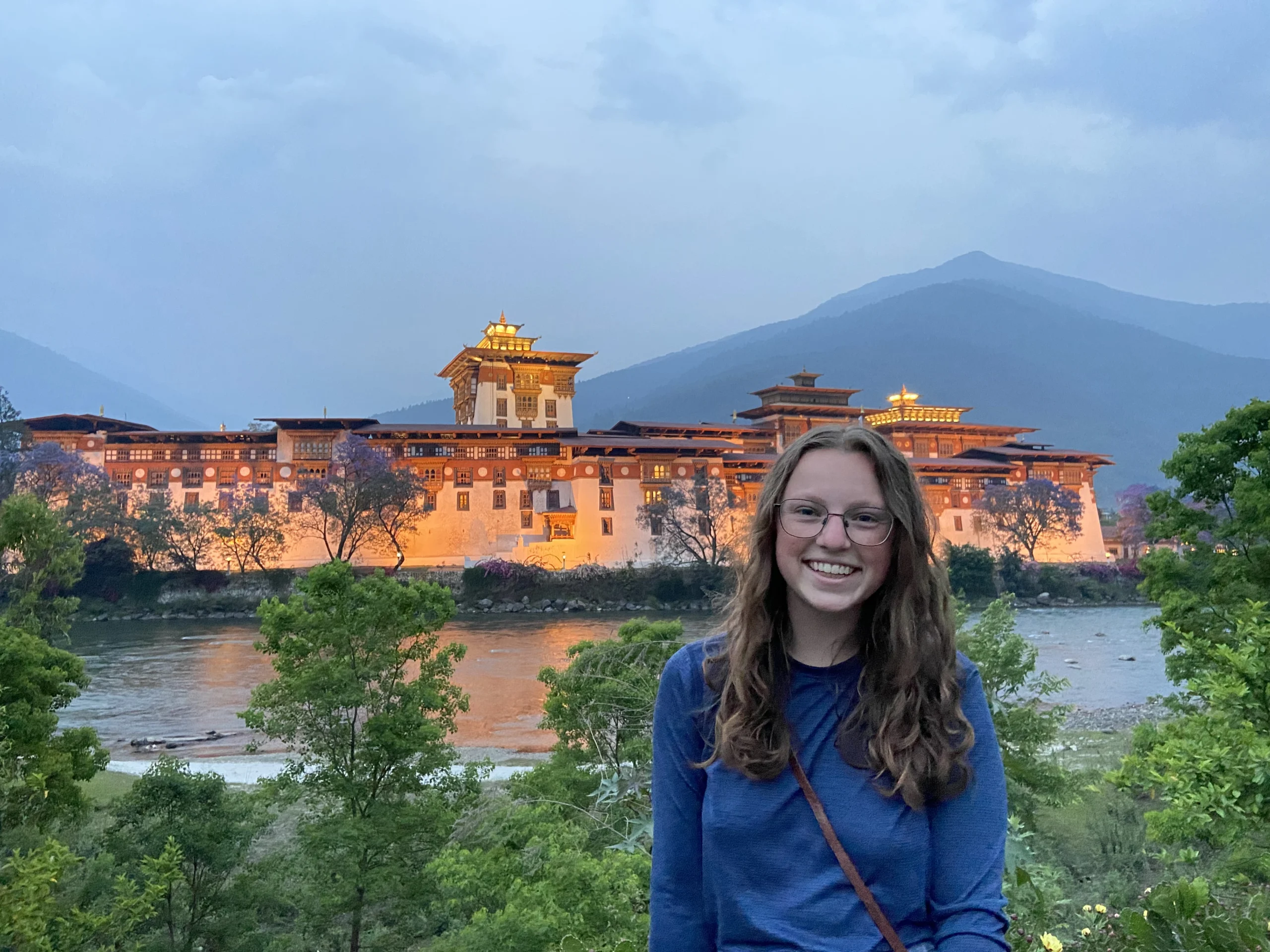 A young person in a blue shirt and glasses standing in front of an asian architectural building across a river