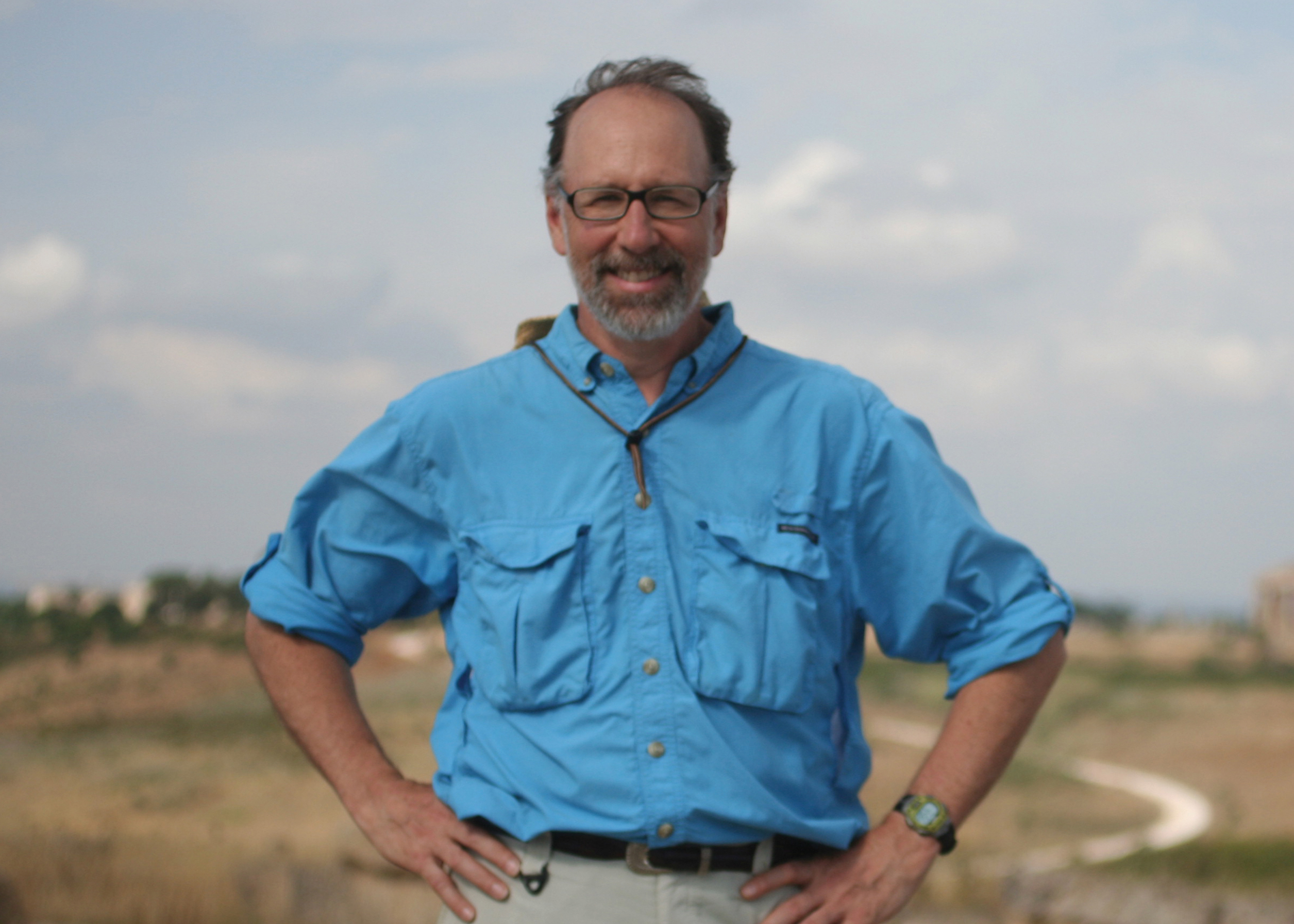 A older man in a blue shirt, glasses, and hands on his hips smiles at the camera