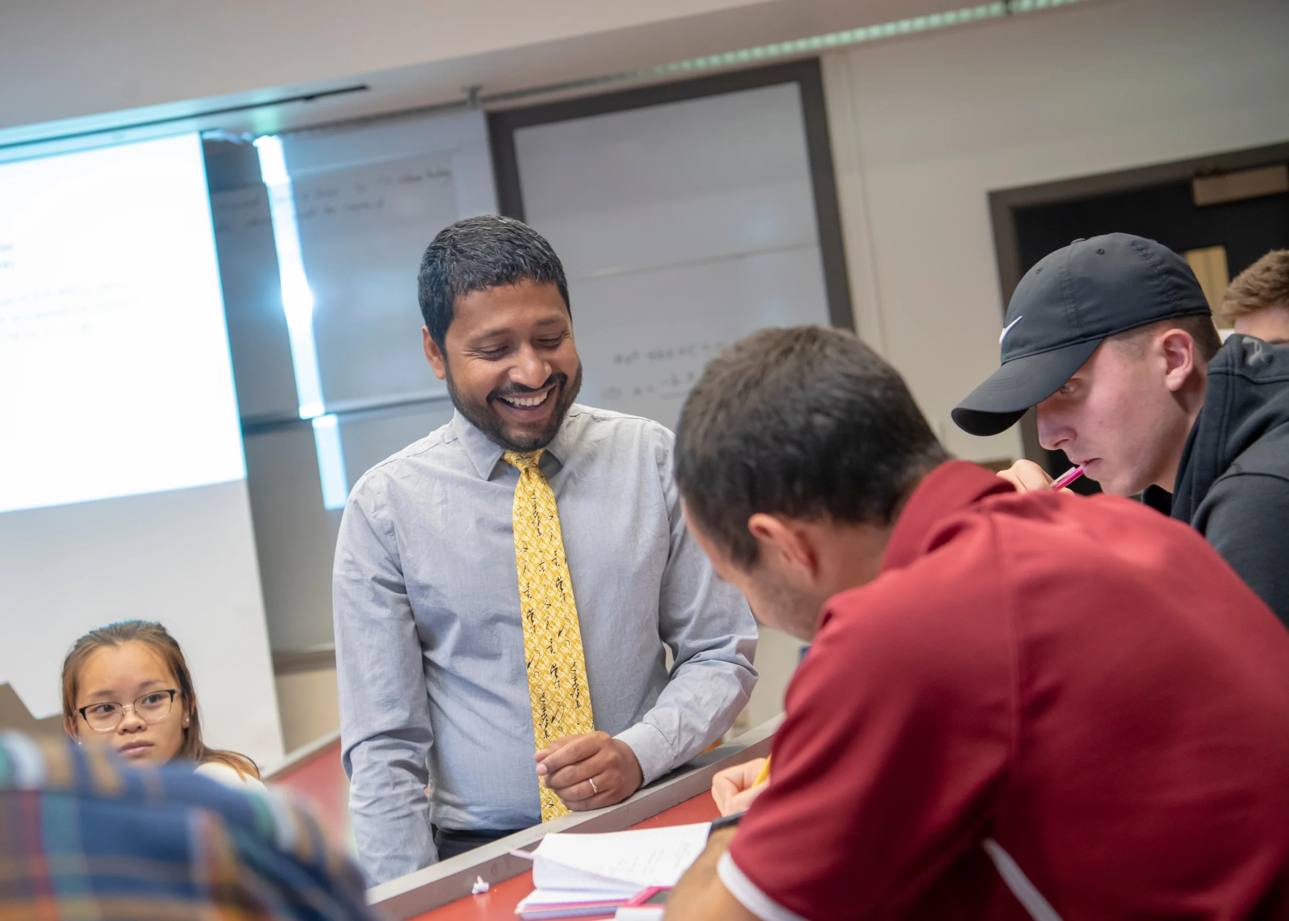 A man in a shirt and tie smiles while speaking with a group of young students in a classroom setting.