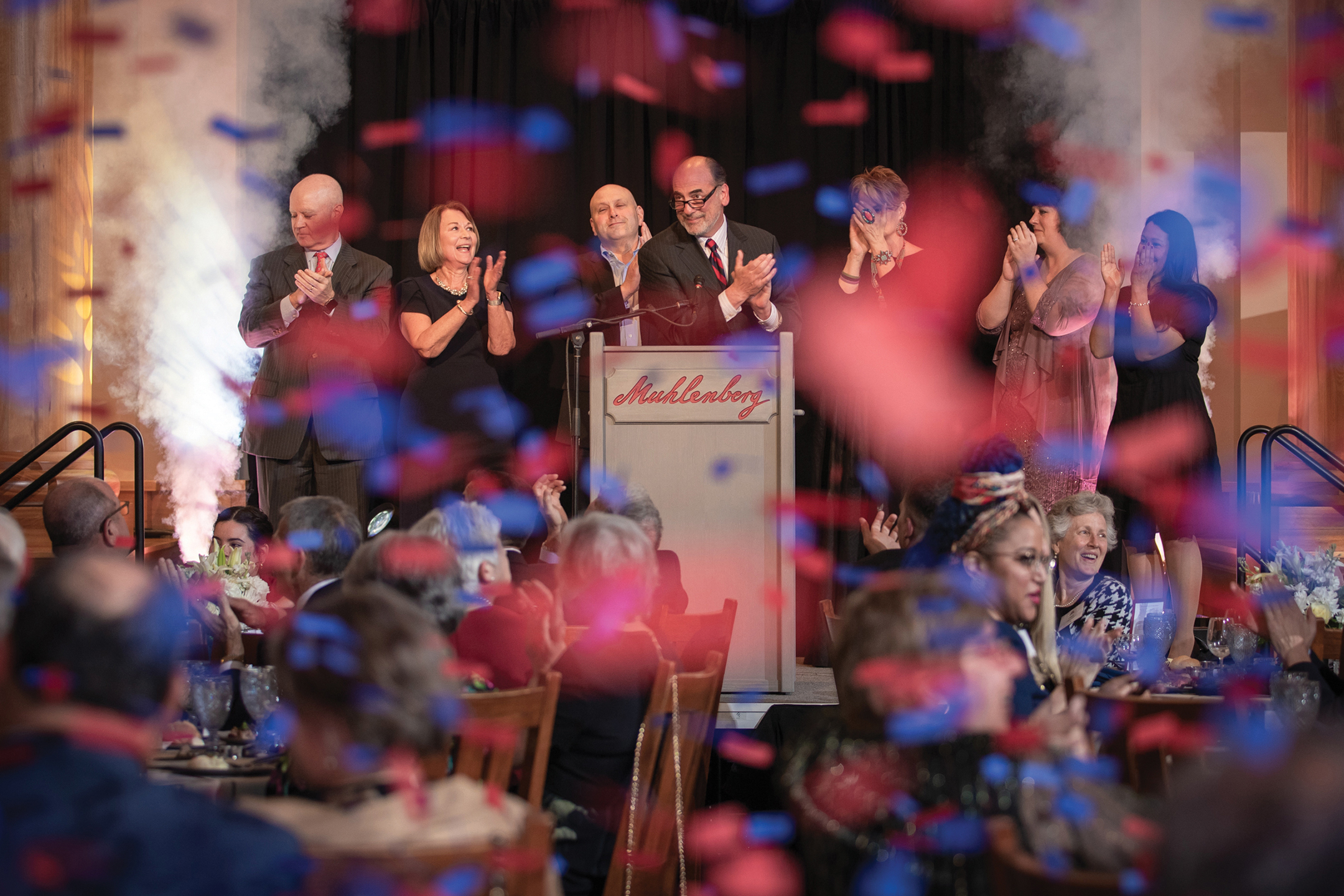 Red and blue confetti falls all around a clapping audience while on stage behind a podium that reads Muhlenberg seven people stand clapping