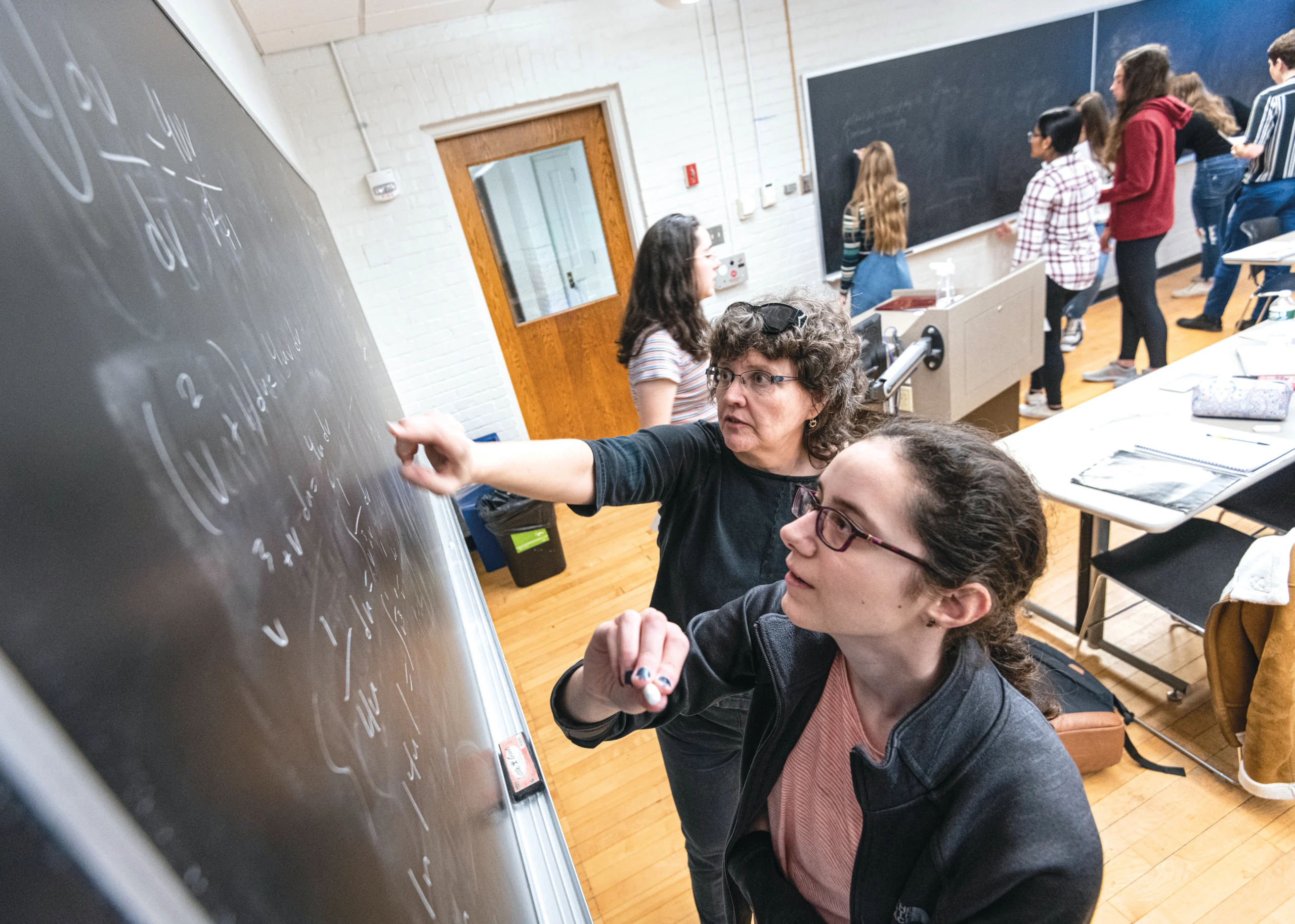 A college professor points to the black board on the left from behind a student in the foreground. More students work in the background.