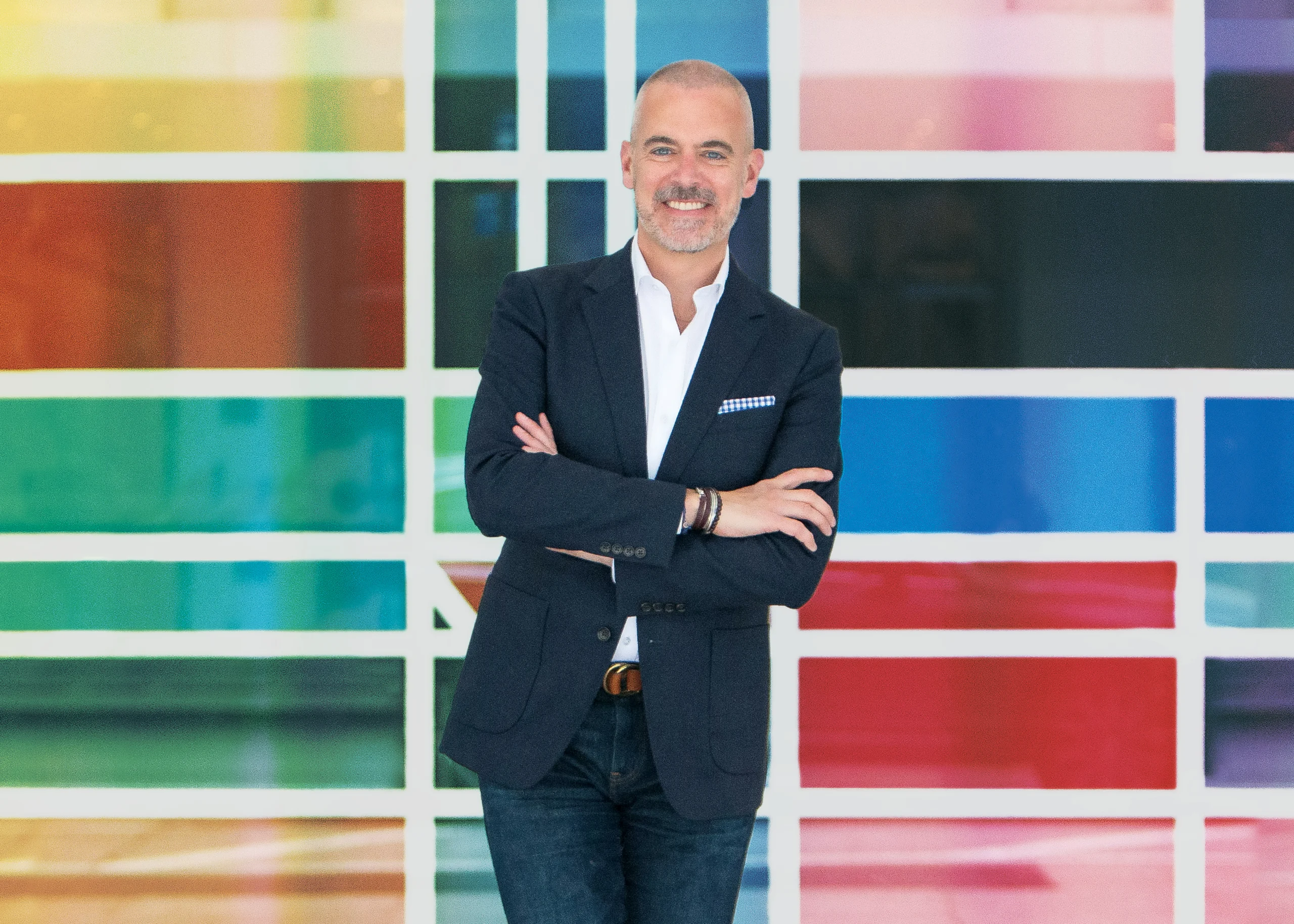 Photo of a man, Mike Doyle, in a suit with his arms crossed and a rainbow colored geometric background.