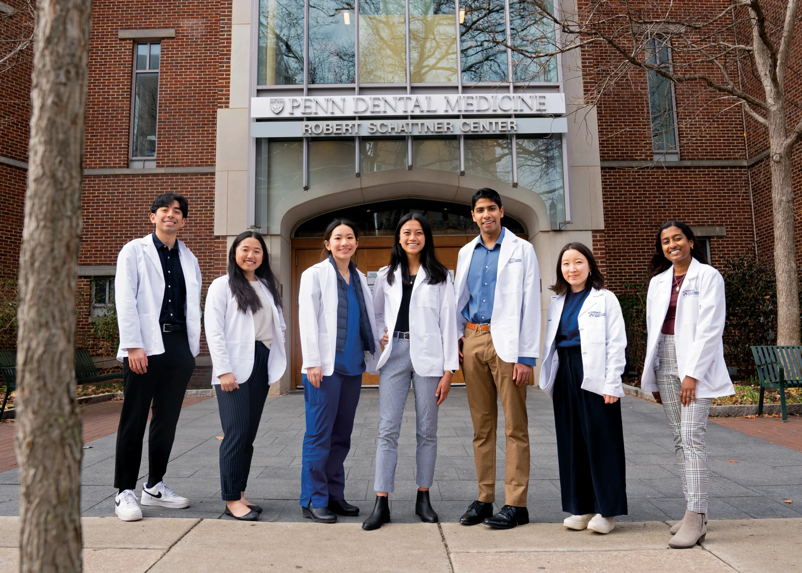 A group of young people lined up in lab coats smiling for the camera in front of a building that says Penn Dental Medicine Robert Schattner Center