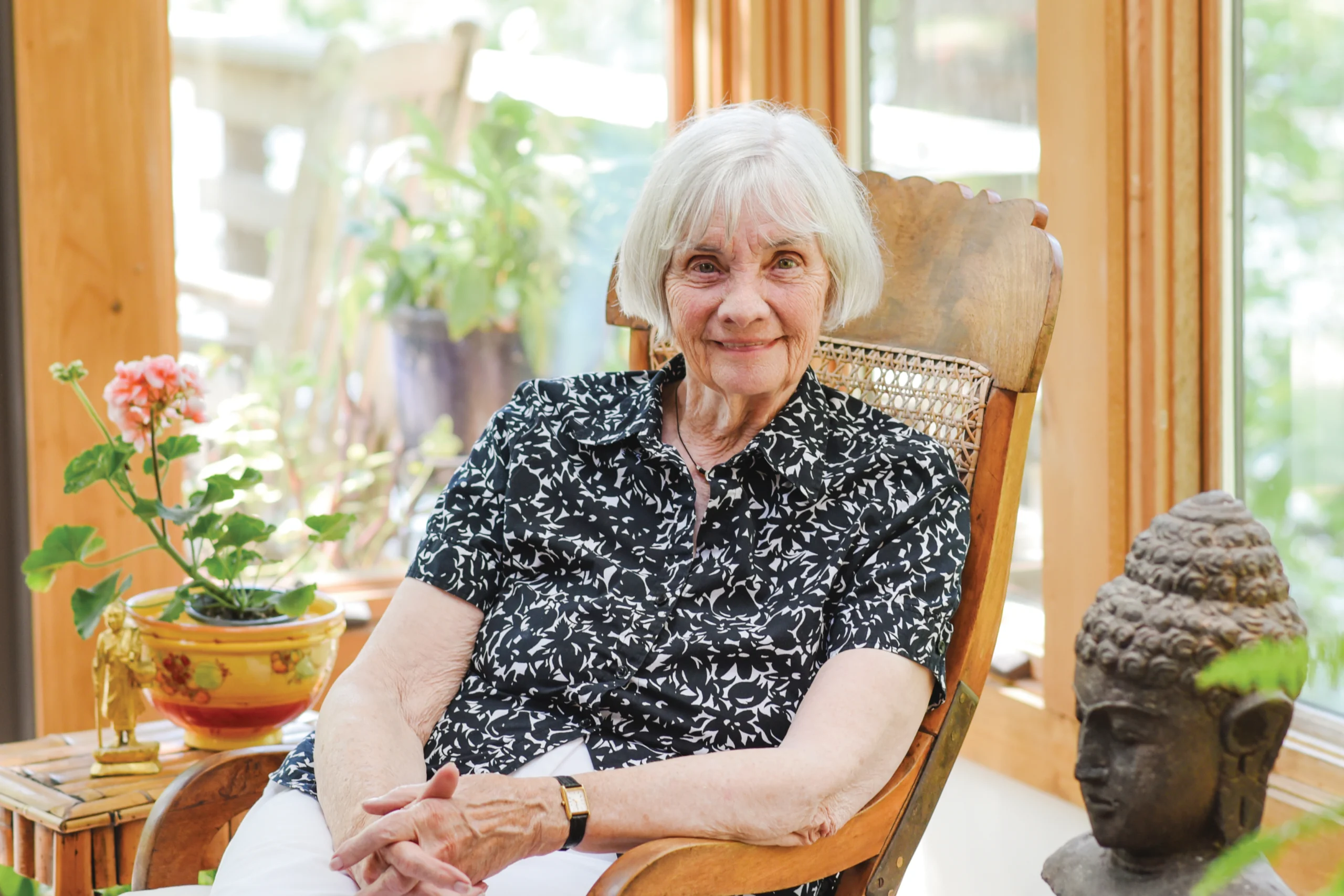 A photo of an older woman in a rocking chair with her hands in her lap and a black and white designed shirt smiling for the camera.