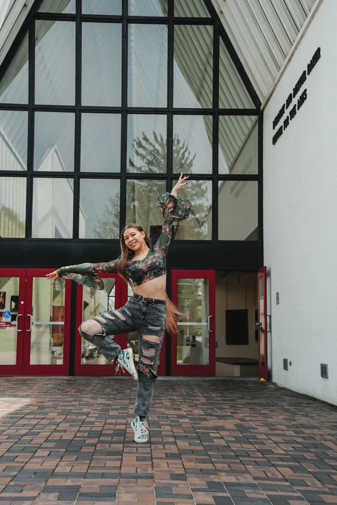 A college student with long hair does a dance pose outside a college building with red doors