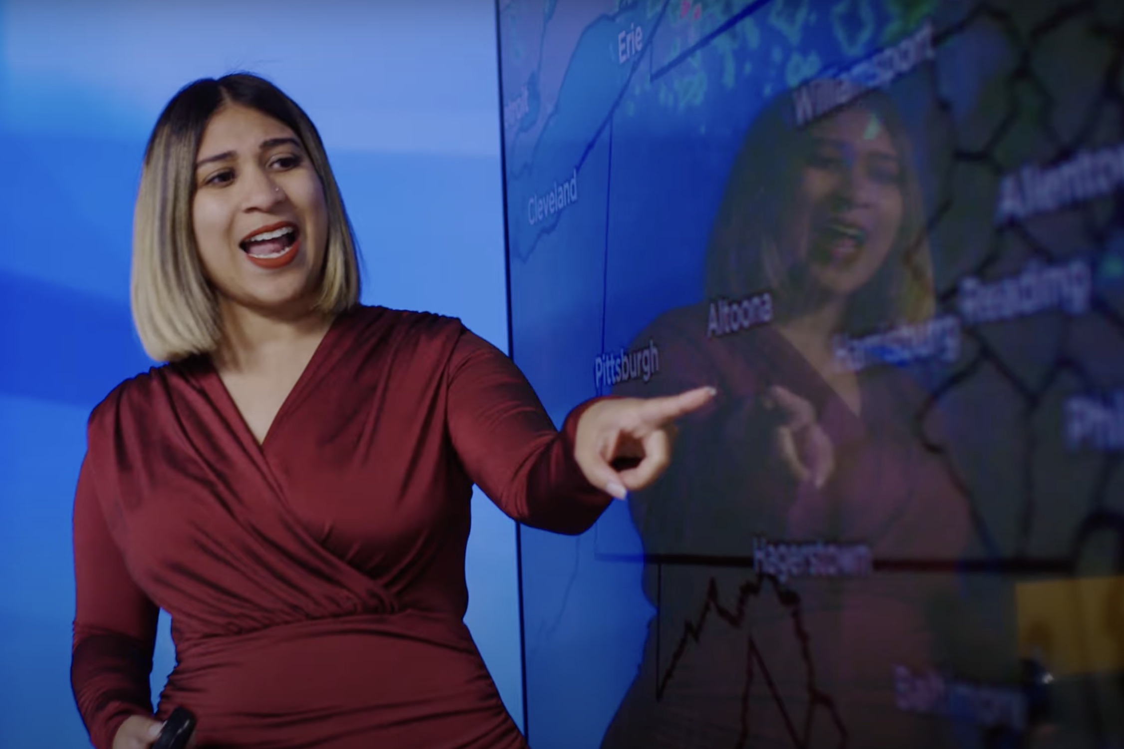 A meteorologist in a red dress points at a screen showing a map