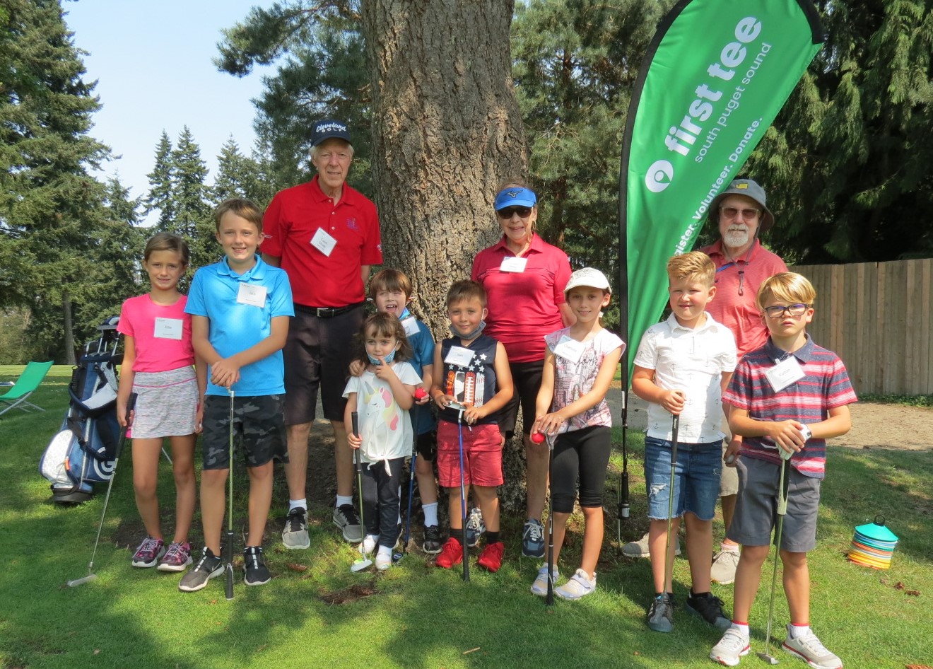 A group of kids with golf clubs with three adults in golf wear and a sign that says "first tee"