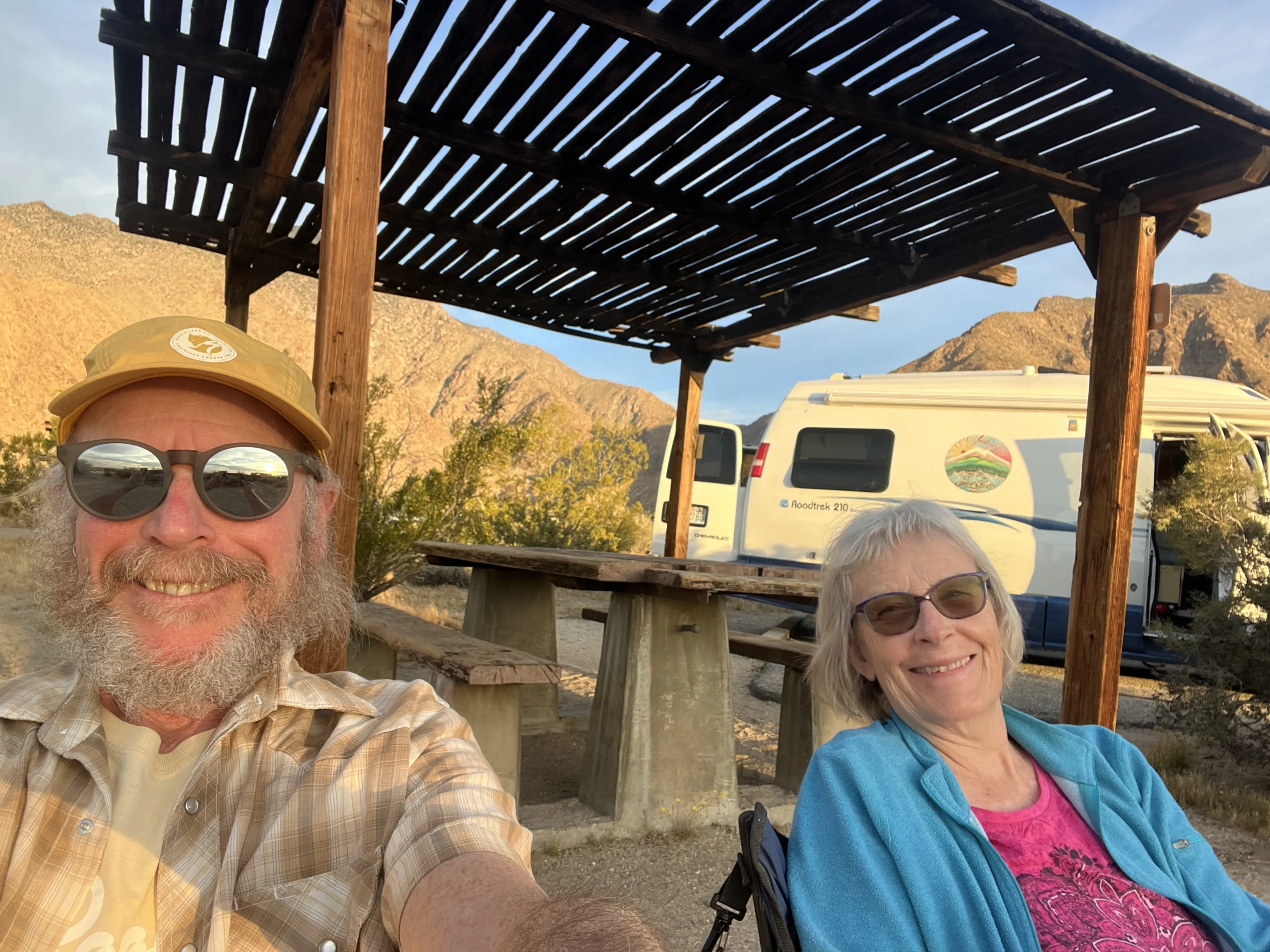 Two adults smile with a van in the background sitting outside in a rocky dry environment