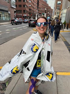 A woman in sunglasses smiles while holding a Boston Marathon blanket on herself