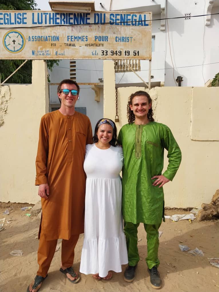 Three young adults in front of a sign that says "LUTHERIENNE DU SENEGAL"