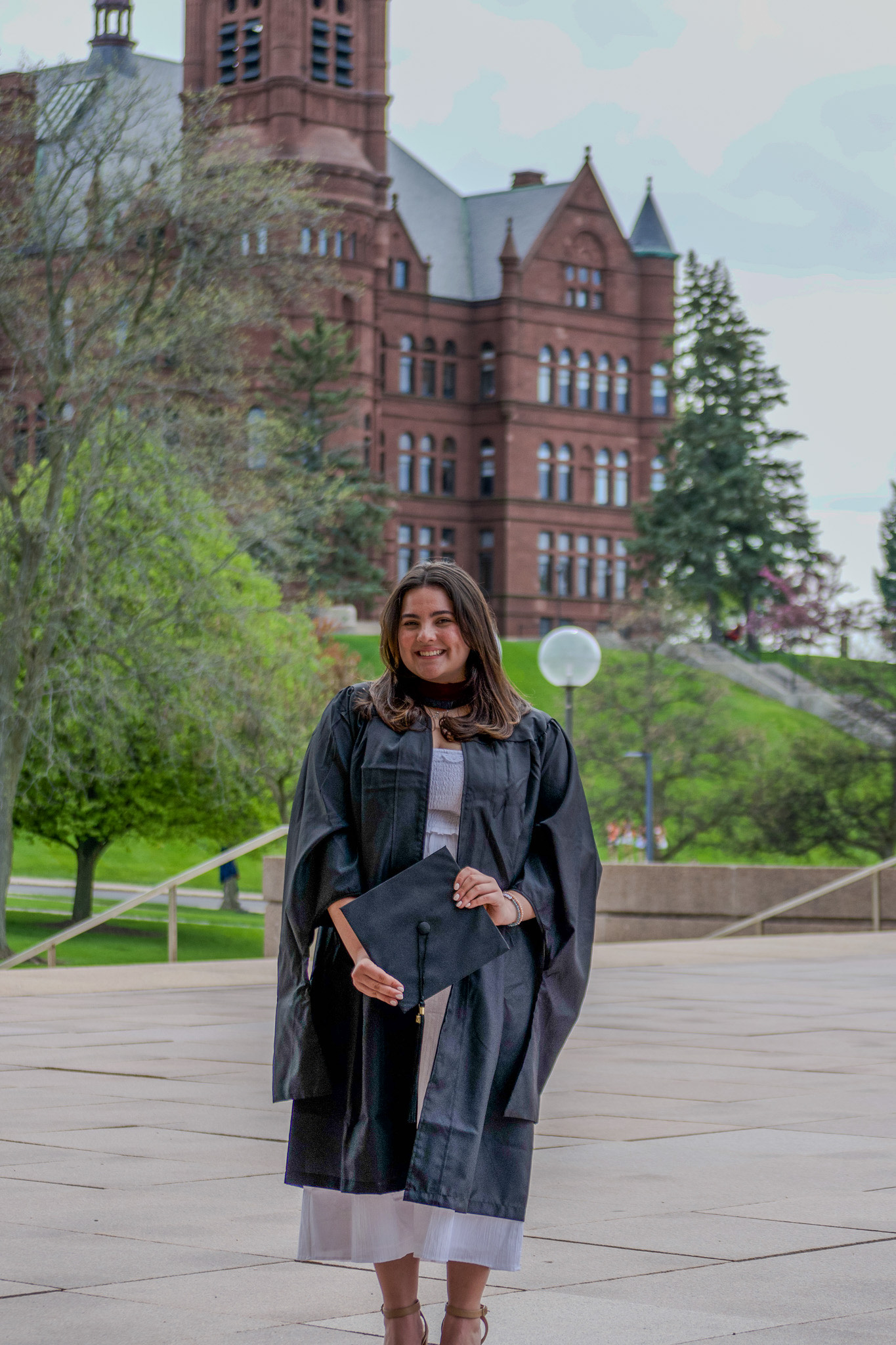 A young adult in graduation attire poses for a photo in front of an old building on a college campus