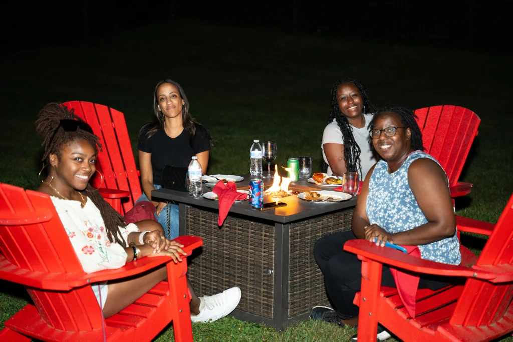 Four women sitting in red Adirondack chairs around a fire pit smile for a photo